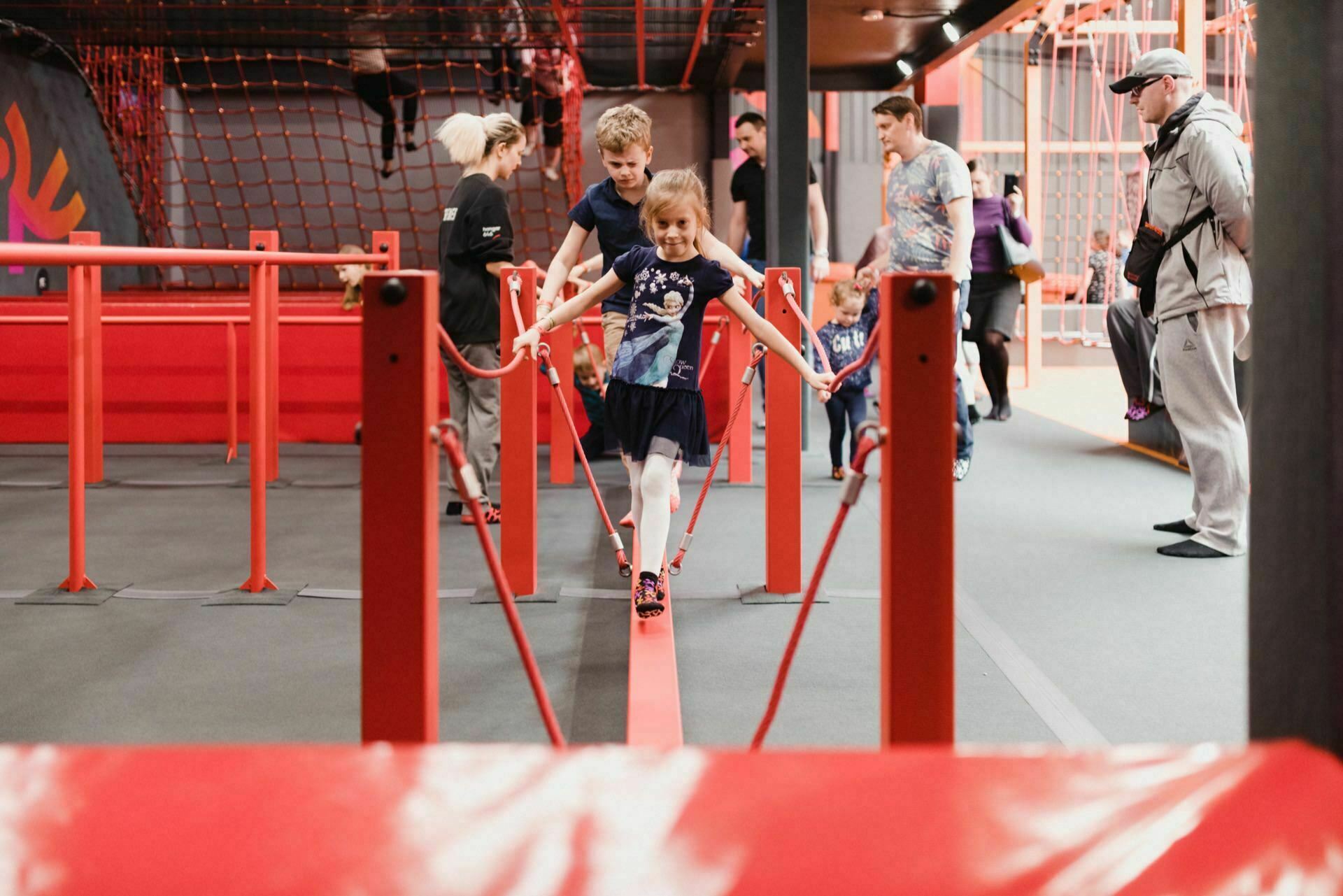 A young girl balances on a red beam in a play center room, surrounded by other people, including a boy and several adults. The environment boasts red and gray structures, nets and gym-like equipment. The lively atmosphere feels as if someone has a *photograph on the wall of the future*, capturing every joyful moment.  