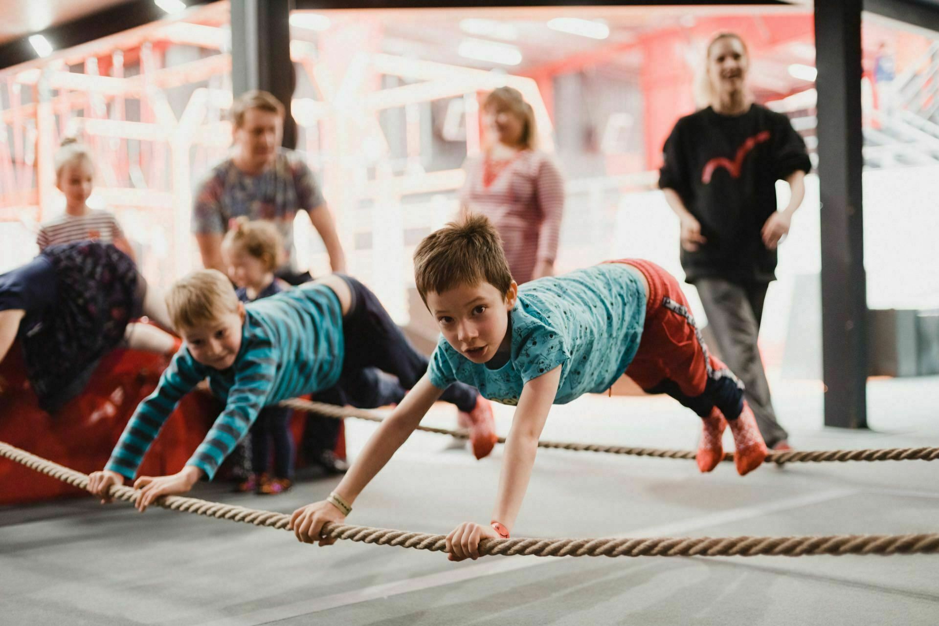 Children play on rope obstacles in an indoor playground. A boy in a blue shirt and red pants balances on the ropes, joined by another boy in a blue striped shirt. Adults stand nearby, smiling and watching the children during the activity, which was beautifully captured by event photography.  