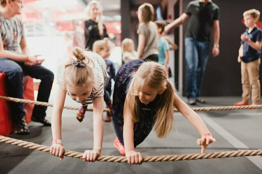 Two young girls crawl under thick ropes during an indoor activity, with other children and adults in the background, captured in a lively group setting by fotografia eventowa Warszawa.