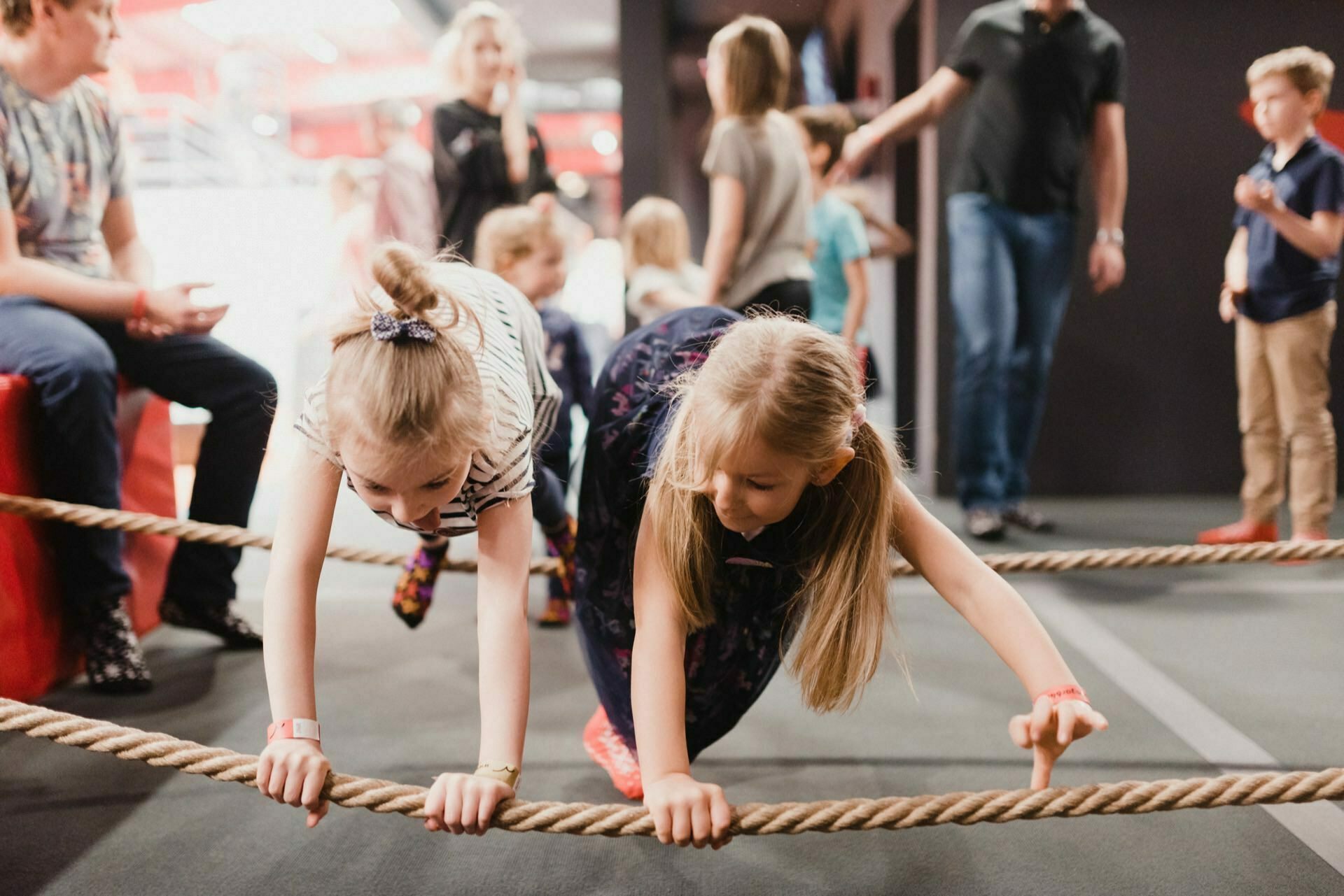 Two young girls crawl under a low rope during an indoor activity. Other children and adults are scattered in the background, making the environment come alive like a typical event photo essay. Several people are watching, and one adult is leading a child around what looks like a recreational area or playground.  