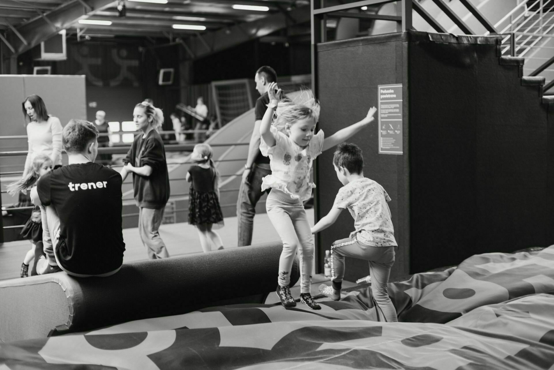 Black and white photo of children vigorously jumping on a padded obstacle in an indoor playground. Sitting nearby is an adult wearing a T-shirt that reads "Coach" (trainer). Other adults and children engage in various activities in the background, turning up the lively atmosphere - event photography capturing pure joy.  