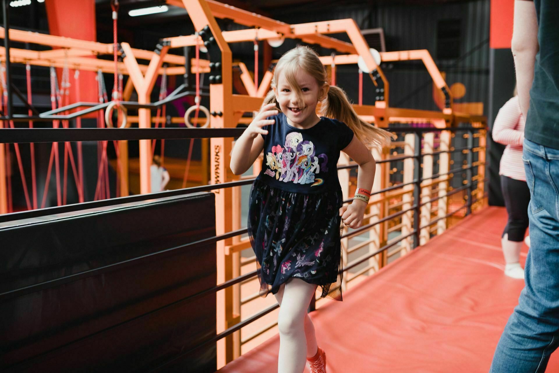 A young girl with blonde hair pinned up in braids, wearing a black dress and white leggings, is running and smiling across the red floor. She is on an indoor obstacle course with orange posts and rings in the background. To the right, an adult's torso and arm are partially visible, providing the perfect moment for event photography.  