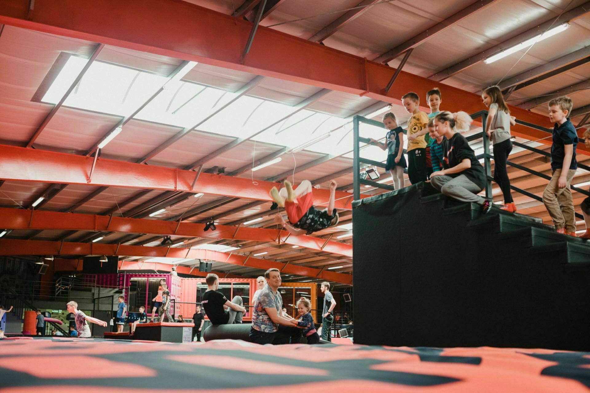 A group of children watch someone perform a back flip in a pool of foam at an indoor trampoline park. The area is illuminated by overhead lights, with various trampolines and play areas visible in the background, and other people enjoying the activity, creating the perfect scene for event photos. 