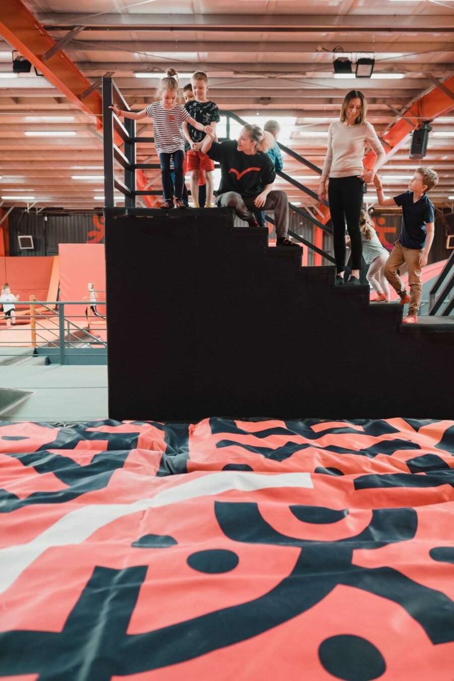 Grupa dzieci i dorosłych stoi i siedzi na klatce schodowej wewnątrz krytego obiektu rekreacyjnego, prawdopodobnie uchwycona przez fotografa eventowego Warszawa. Wygląda na to, że dyskutują lub przygotowują się do jakiegoś działania. Na pierwszym planie duża, kolorowa powierzchnia z geometrycznymi wzorami.