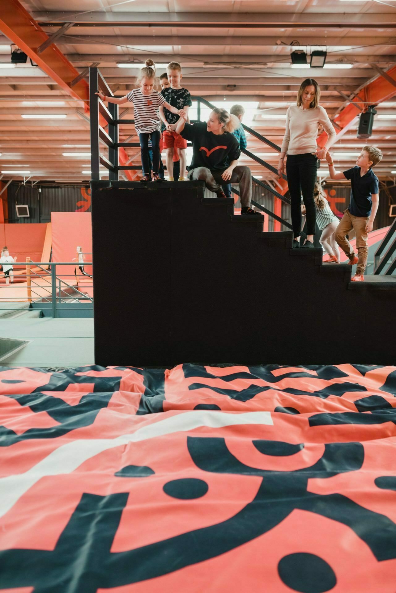 A group of children and adults are standing and sitting on a staircase inside an indoor recreational facility, probably captured by an event photographer Warsaw. They appear to be discussing or preparing for some activity. In the foreground is a large, colorful area with geometric patterns.  