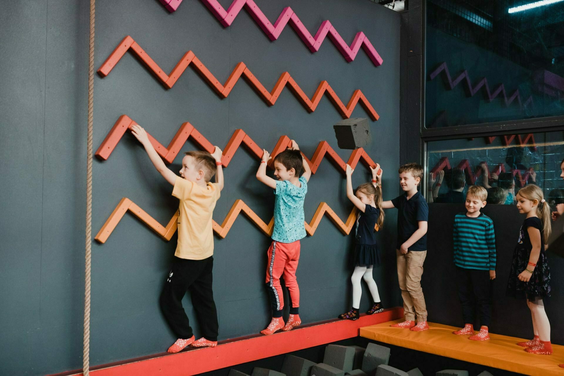 Children enjoy indoor activities. They climb the zigzag-shaped wall, most using the wall handholds, while others wait their turn. The wall has pink, orange and red handles, and the children are wearing colorful outfits and orange climbing shoes. The *birthday photo* captures their joy.   