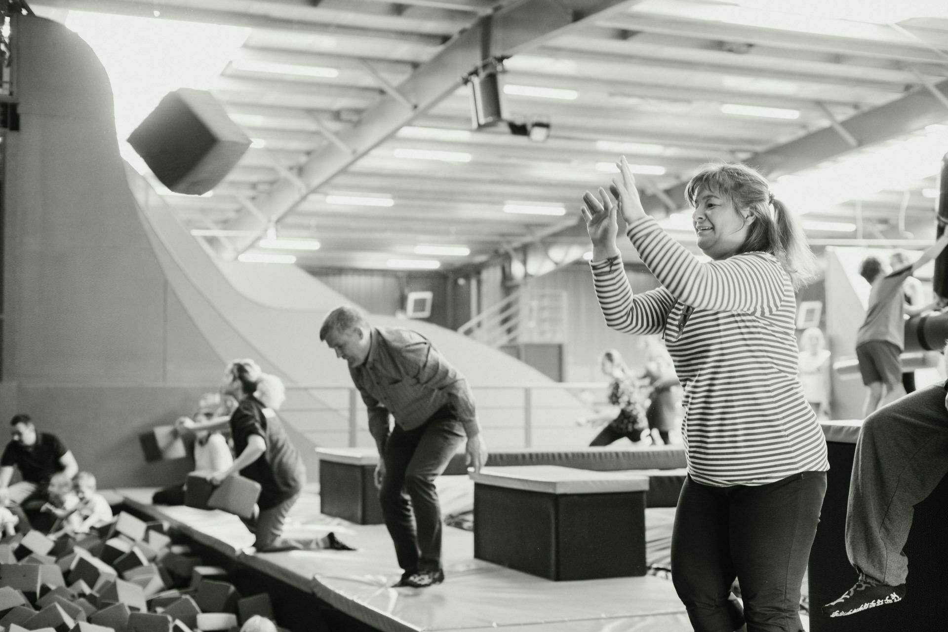 Black and white event photo of people in an indoor trampoline park. A woman in a striped shirt catches a foam block, others in casual clothes are actively engaged in games. Scattered throughout the event photo are foam blocks with happy expressions on their faces.  