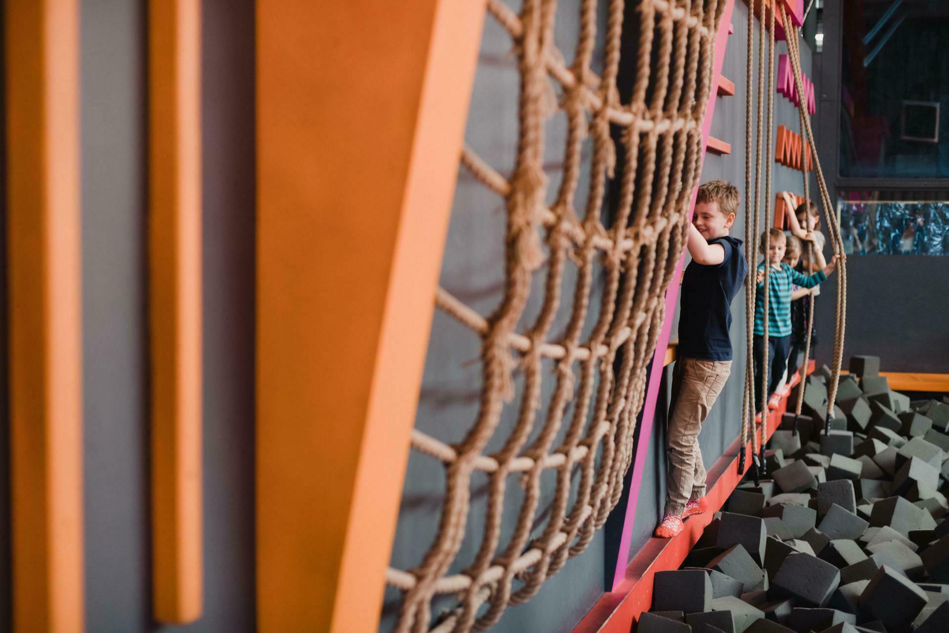 Two children play in an indoor adventure park. They cover a route using a rope net and balance beam, surrounded by foam blocks. The child in the foreground supports the net by balancing on a beam, as beautifully captured in this scene from the event.  