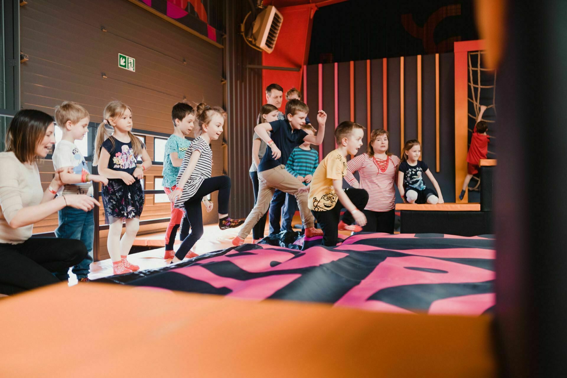 Children and adults enthusiastically line up and prepare to jump into the colorful, padded playground of the indoor recreation facility. The room is decorated in bright colors, which perfectly reflects the fun and energetic environment, perfect for photo coverage of the events. It looks like everyone is having a great time.  