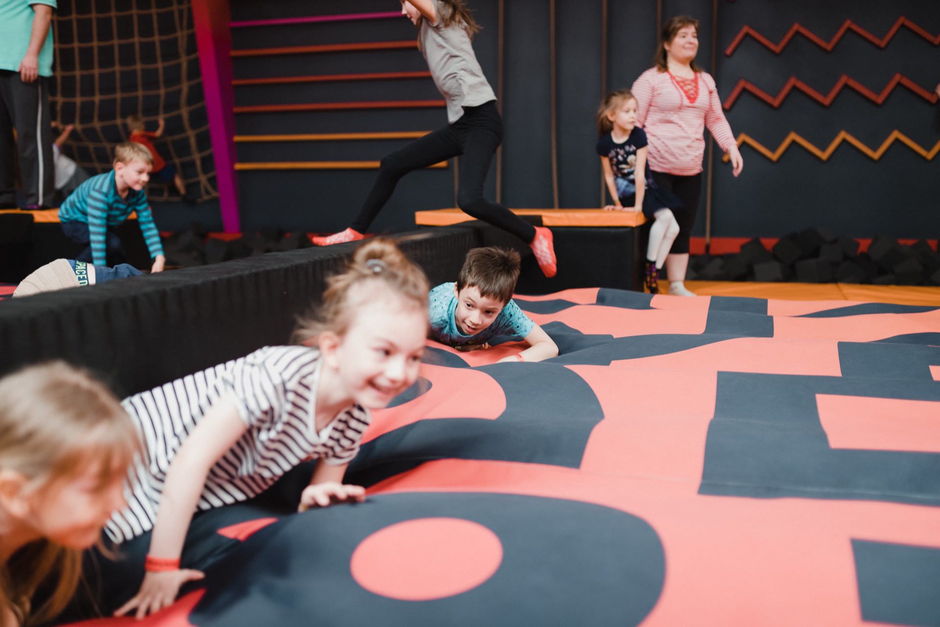 A group of children are energetically playing on an indoor playground. They climb and crawl on the cushioned play equipment. The environment is colorful and vibrant, with one woman supervising them in the background, perfect for an event photo shoot to capture joyful moments.  