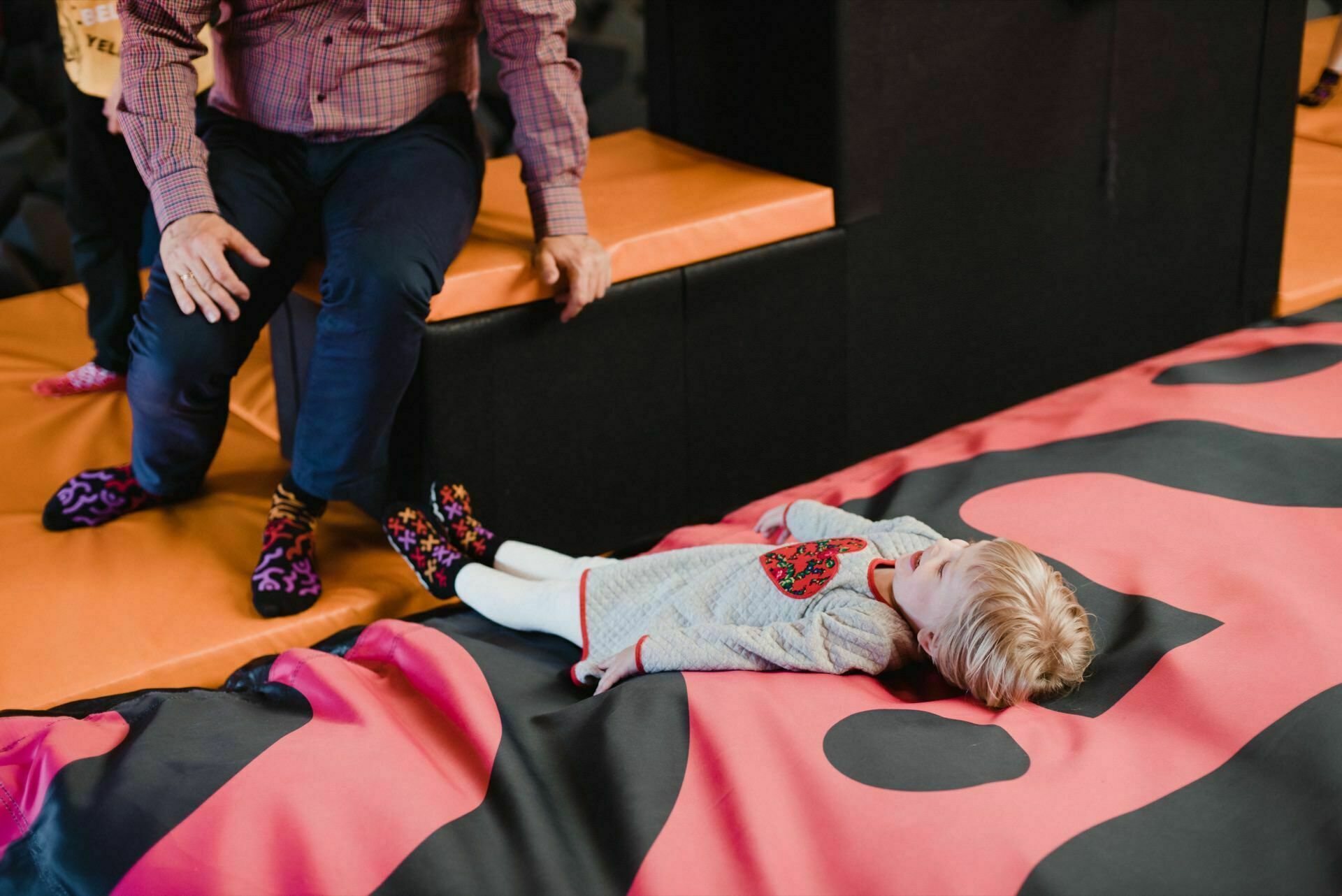 A child dressed in a white top with a red pattern lies on his back on a large, soft play mat. Nearby sits an adult, dressed in a checkered shirt and dark pants with colorful socks, watching and extending his hand toward the child. In the background is padded play equipment. This is the perfect moment for *event photography*.   