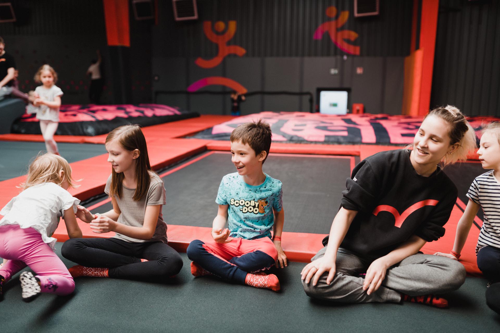 A group of children and a young woman sit on the floor of an indoor trampoline park. They are smiling and having a fun conversation, which is perfectly captured in this event photography. In the background, trampolines and colorful wall graphics create a lively event photo.  