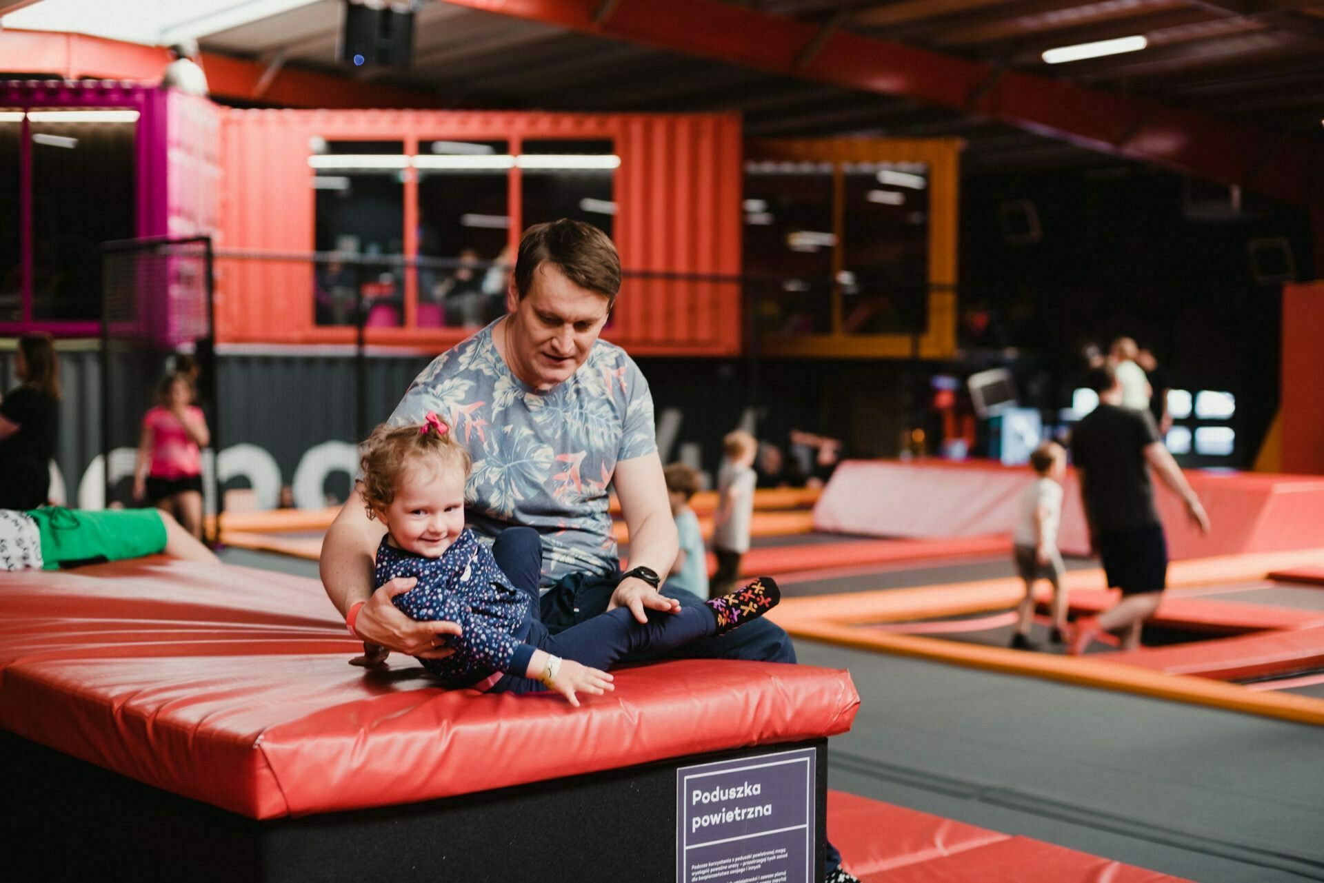 A man helps a young child slide off a padded platform at an indoor trampoline park. Other people and children can be seen in the background doing various activities on the trampolines and play structures, capturing the perfect moment for any photographic event. 