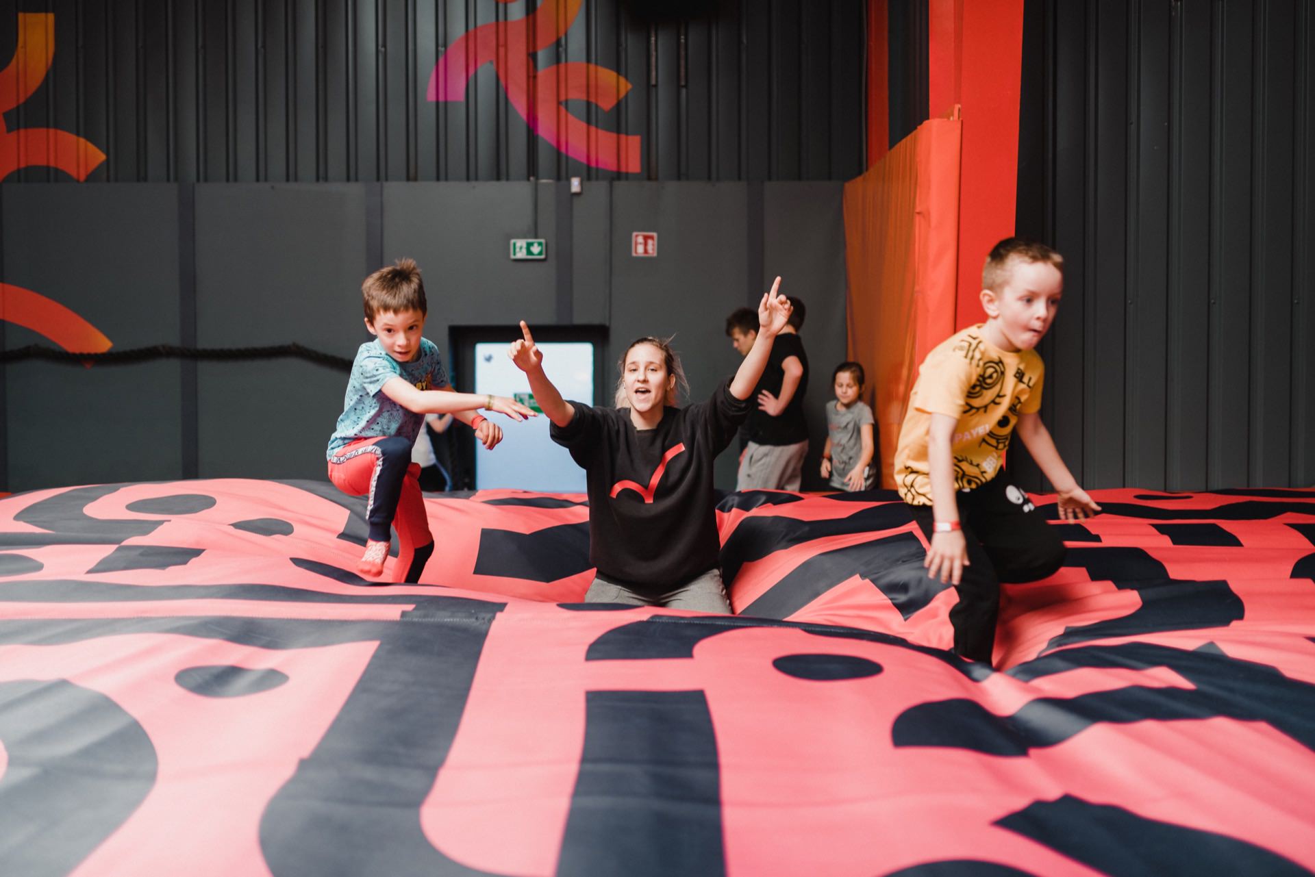 A group of children play on an inflatable area with colorful patterns. One woman kneels in the middle, smiles and points upward. A photographer on a family platform captures the joyous moment as two boys on either side enthusiastically climb and jump on the inflatable surface.  