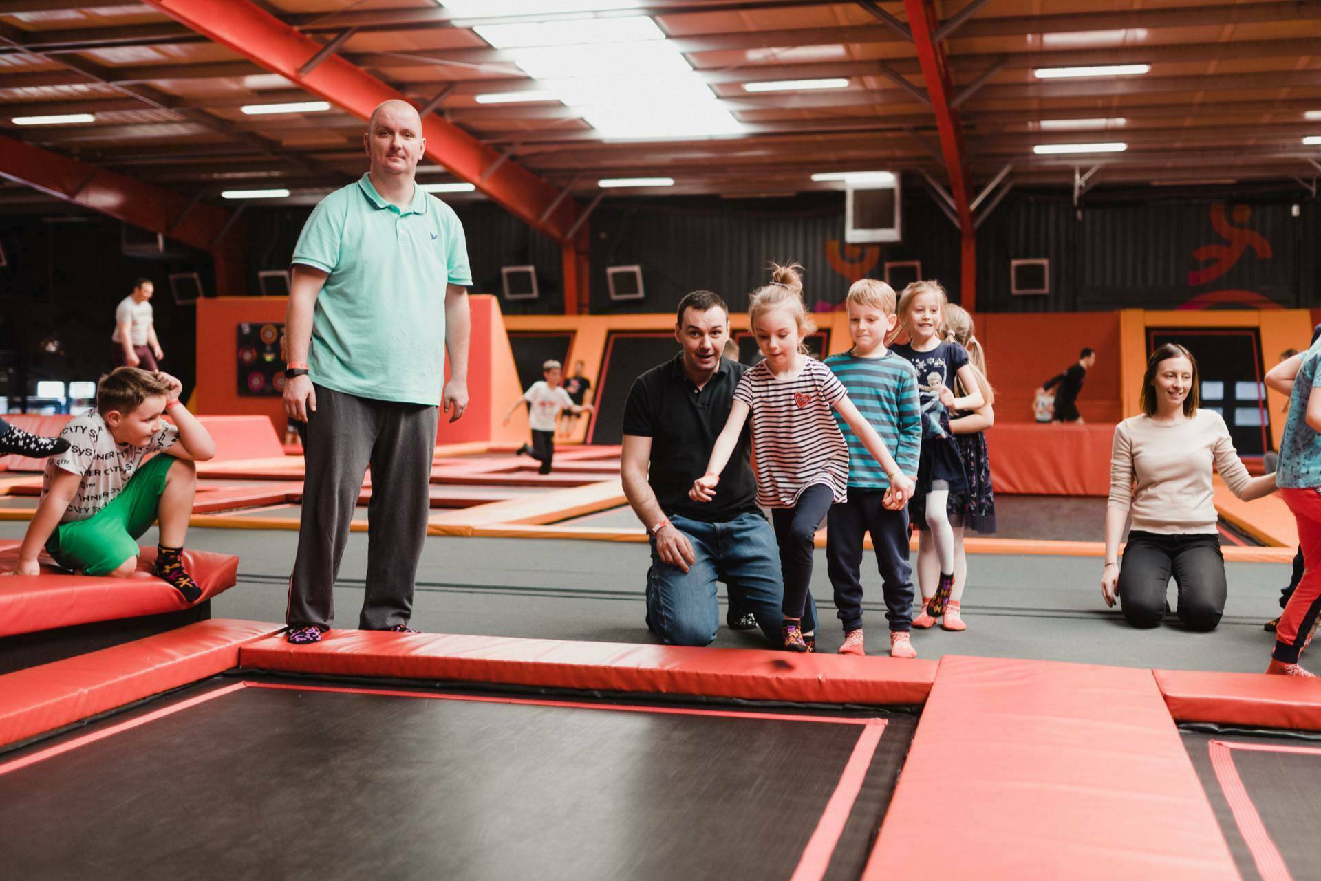 A group of people, including children and adults, are having fun in an indoor trampoline park. The children stand on the edge of the trampoline, ready to jump, while the adults are nearby, supervising and smiling. The vibrant and spacious environment is perfect for a heartfelt photo-op of the events.  