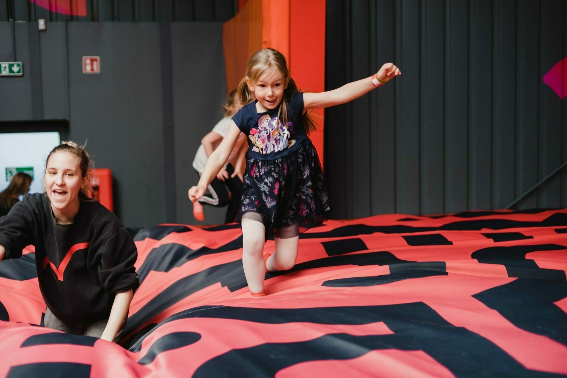 A young girl balances, walking on a large, pink, inflated play structure. Nearby kneels an adult woman in a black sweater with a red heart motif, smiling and supporting the girl. In the background are dark walls and other exercise equipment - perfect moments captured for event photographer Warsaw.  