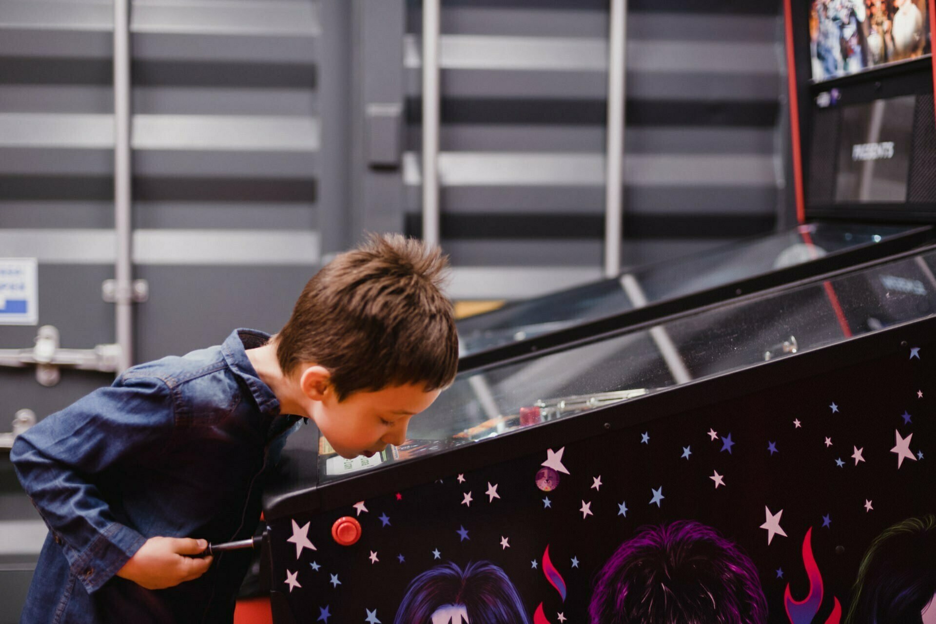 A young boy in a denim shirt looks intently at a flipper playing field with colorful graphics. He leans in to get a better look at the details while holding the side of the machine, creating the perfect moment for event photography. In the background are walls with gray stripes.  