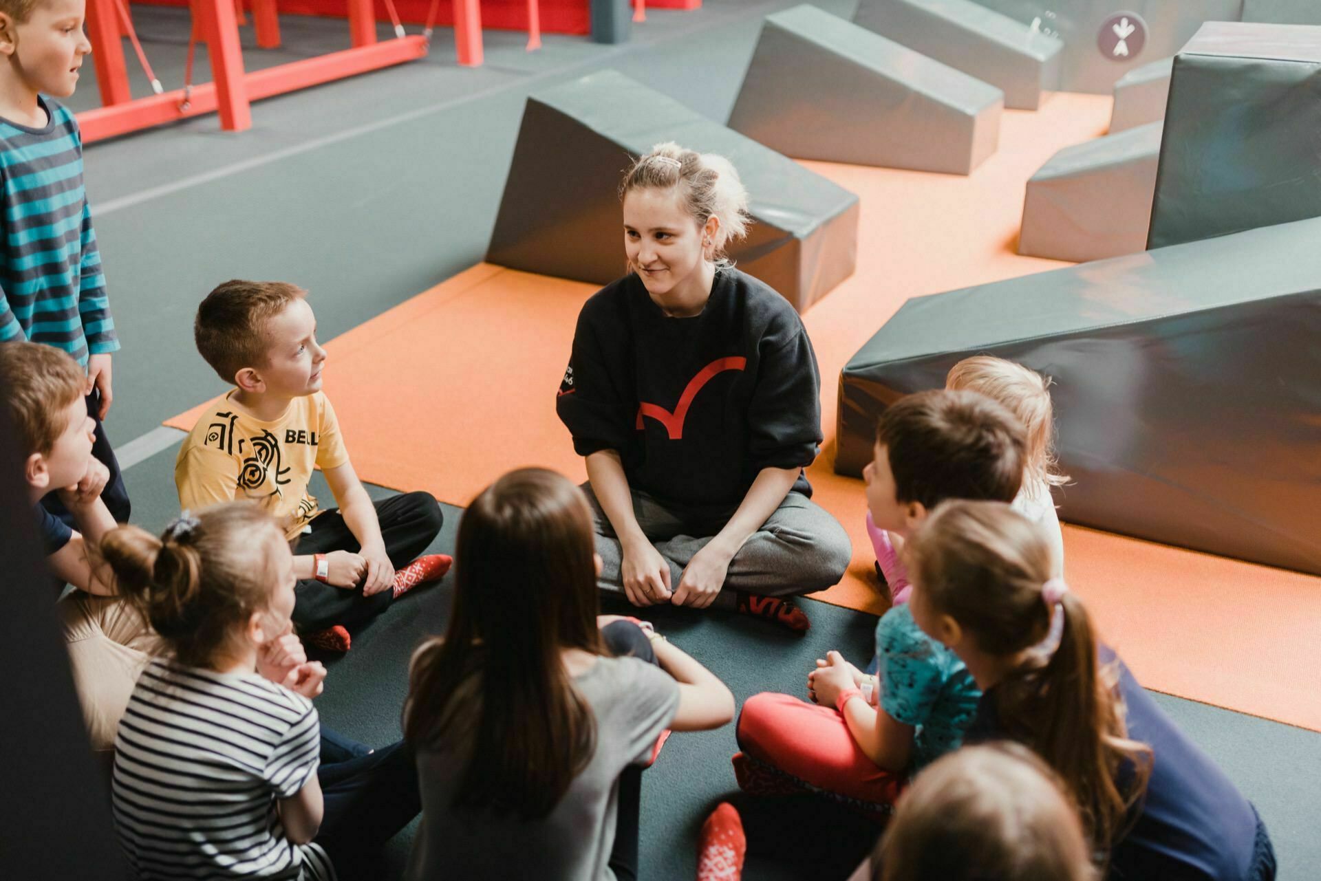 A group of children sit in a semicircle on the floor around an adult woman who smiles and talks to them. They are in what looks like an indoor playground or activity with padded structures and orange mats. A photographer from "Event Photography" captures the attentively listening children.  