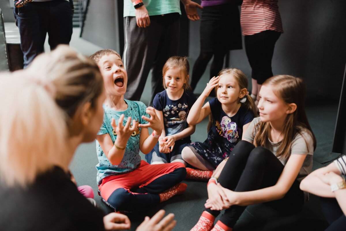 Grupa dzieci siedzi na podłodze w półkolu wokół osoby dorosłej. Jedno z dzieci, ubrane w niebieską koszulę i czerwone spodnie, wygląda na podekscytowane z podniesionymi rękami, podczas gdy inne siedzą i uśmiechają się uważnie. W tle widać inną osobę dorosłą, która rejestruje fotorelację z wydarzeń.