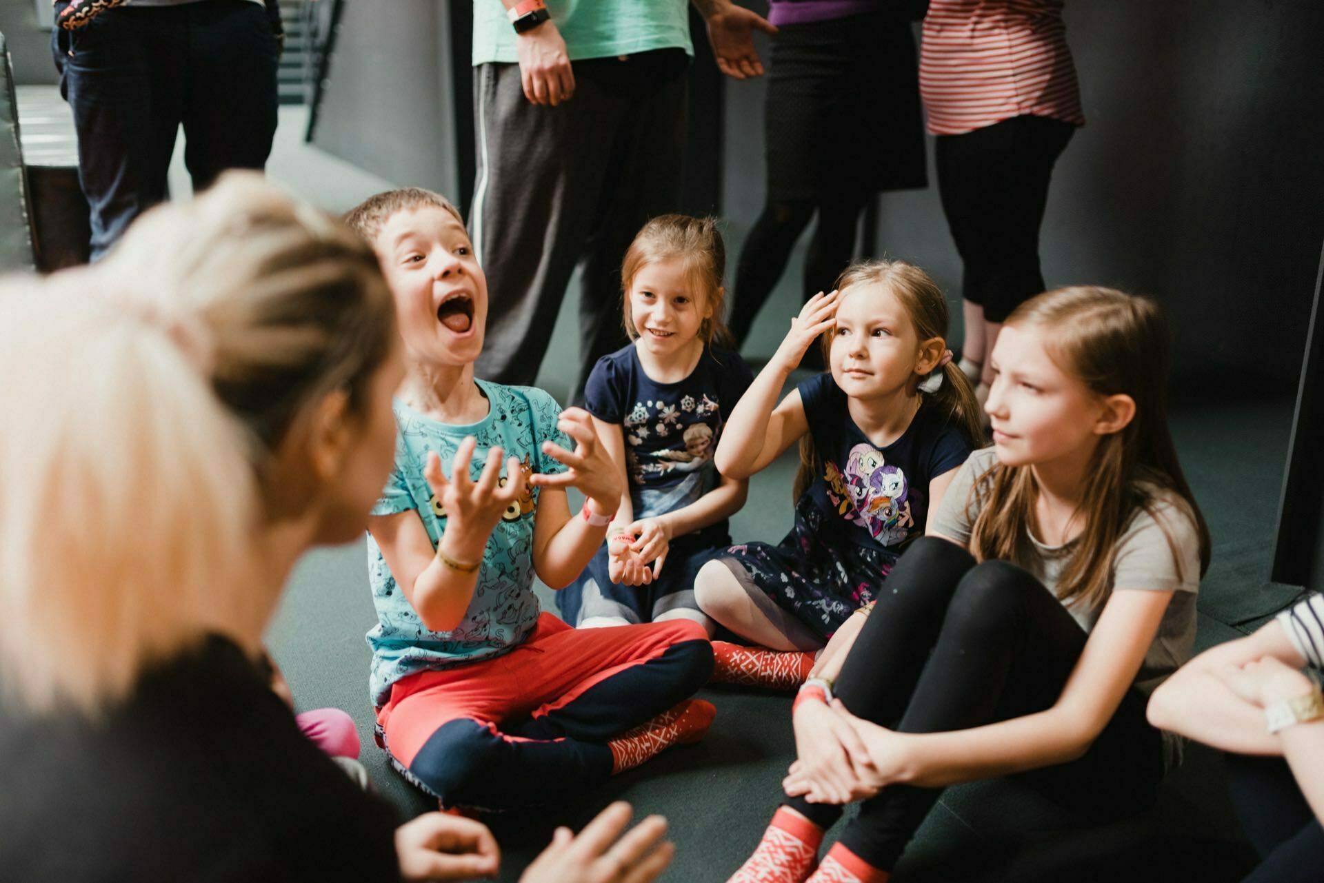 A group of children sit on the floor in a semicircle around an adult. One of the children, wearing a blue shirt and red pants, looks excited with his hands raised, while the others sit and smile attentively. Another adult can be seen in the background capturing a photo report of the events.  