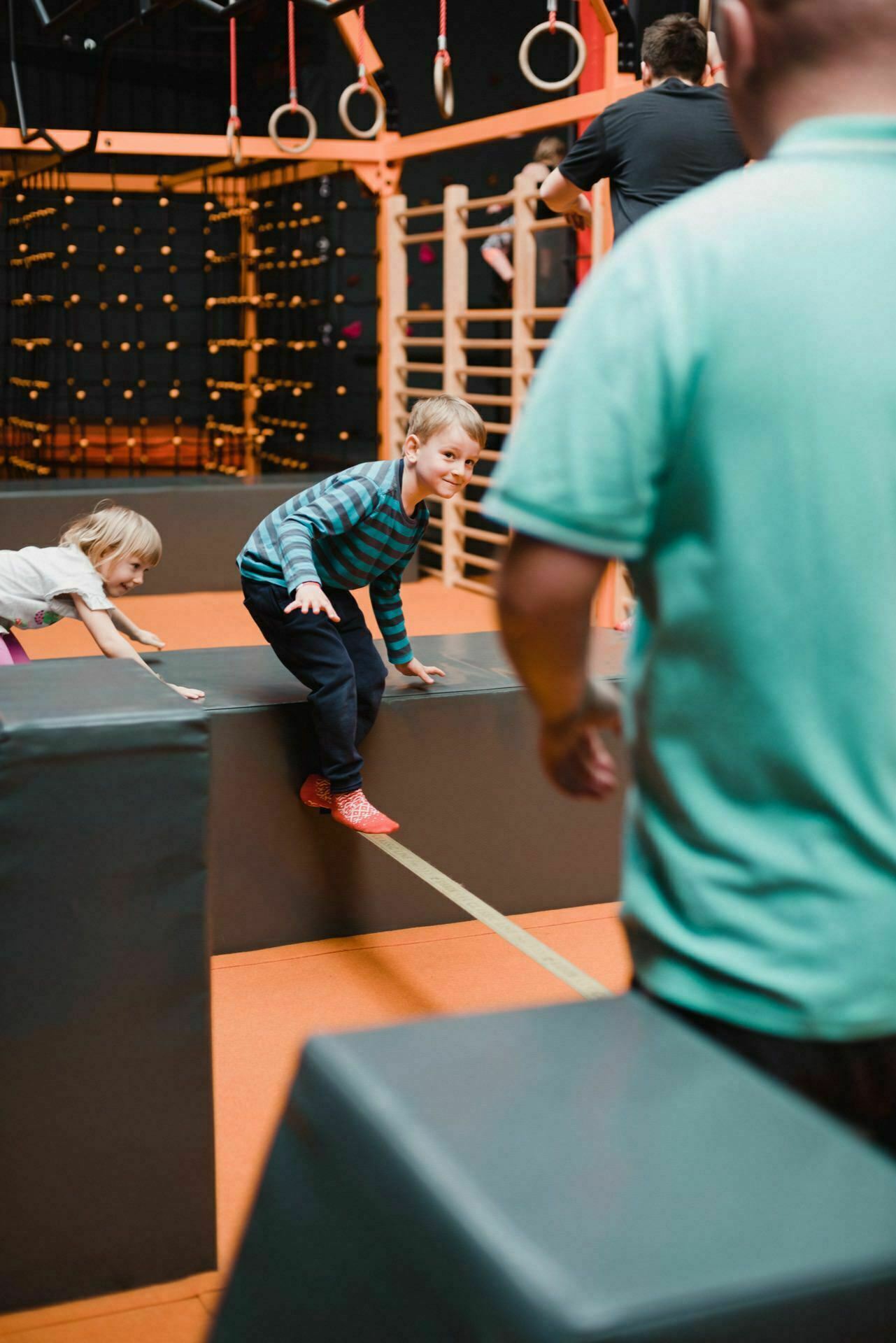 A young boy in a striped shirt and red shoes balances on a beam in a gymnasium, watched by a person in a turquoise shirt. A young girl in a pink shirt and gray pants climbs onto a nearby platform. The gymnasium, ideal for event photography, has climbing structures and rings in the background.  