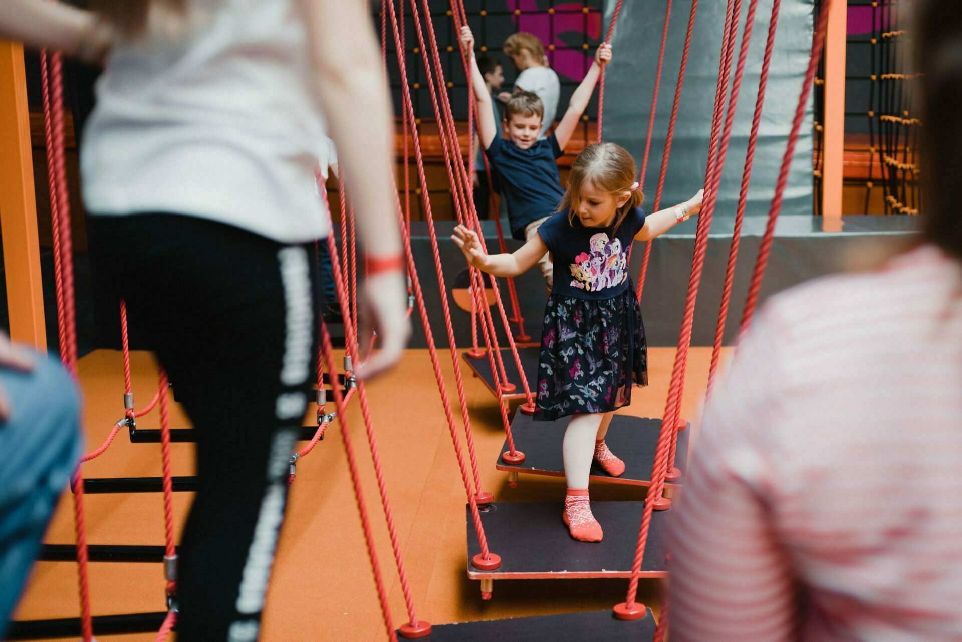 Children play in an indoor playground, moving across a suspended rope bridge using red ropes and black boards. Balancing is a girl in a black dress and orange socks, while behind her a boy holds on to the ropes. Other children can be seen in the background, beautifully captured in this event photo.  