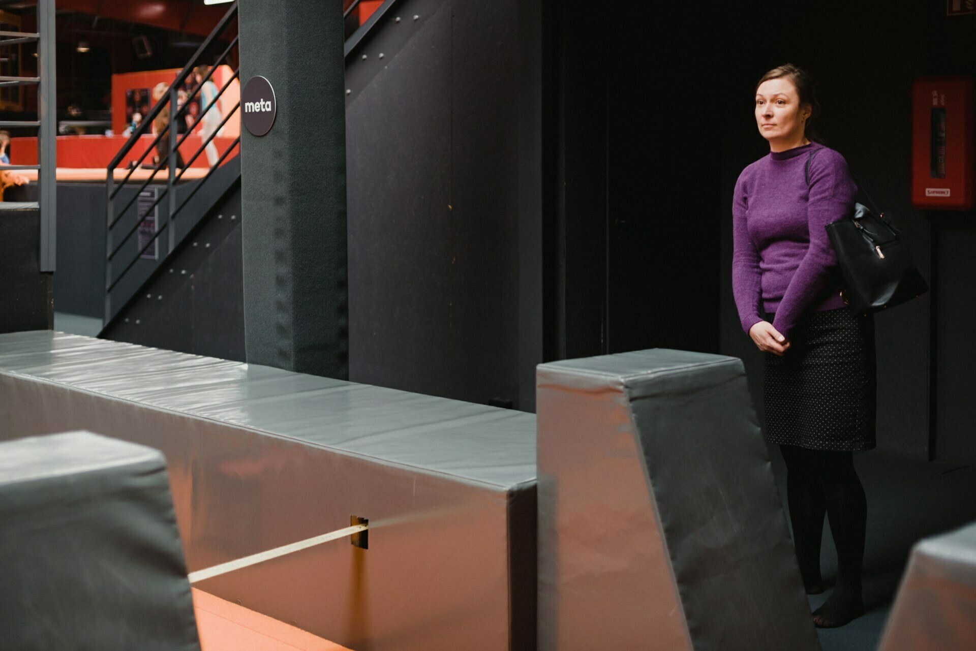 A woman in a purple sweater and black skirt stands by padded exercise equipment in a dimly lit room, typical of many "event photography" shoots. She holds a black bag over her shoulder. In the background you can see a staircase and a black column with a circular sign reading "meta.  