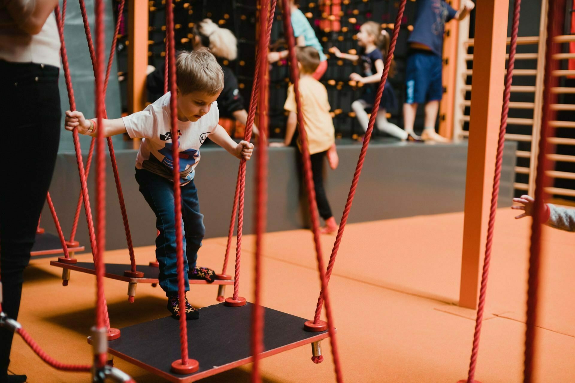 A young boy carefully navigates an obstacle course suspended on red ropes, focusing on his steps. In the background, other children and adults are engaged in various activities, climbing walls and enjoying the indoor playground in an orange and black color scheme - a perfect scene for event photography. 