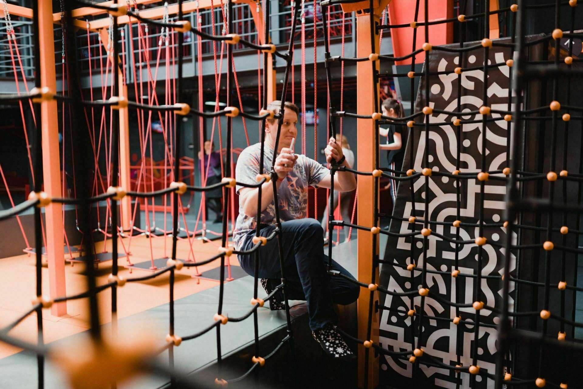 A person in athletic attire navigates a complex rope course in a well-lit enclosed room. They hold the ropes with both hands, maneuvering their feet through the netting, against a backdrop of various climbing elements and a patterned wall, ideal for event photography. 