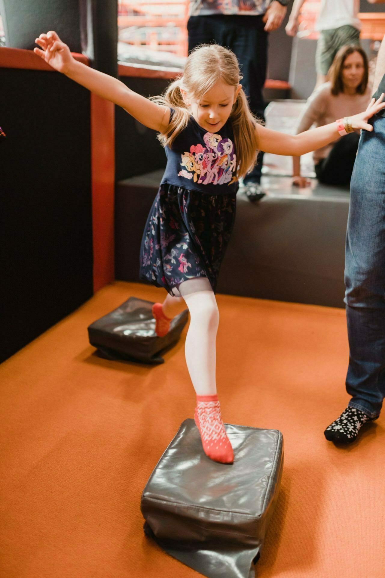 A young girl with blonde hair tied in braids balances on one leg on a padded obstacle during an event photography warsaw. She is dressed in a navy blue dress with colorful characters, white tights and red socks. The floor is orange, with another person partially visible in the background.  