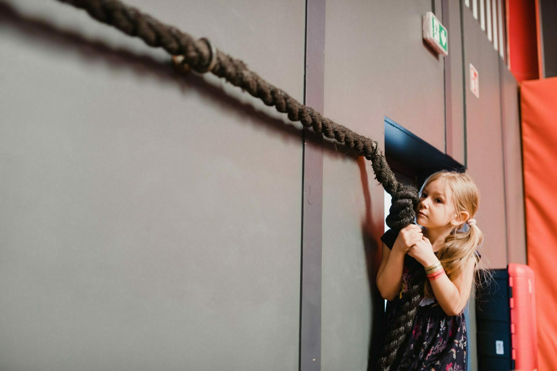 A young girl with braids, wearing a dark dress, climbs a thick rope in a room. She looks determined as she climbs a rope attached to a gray wall. The background contains red and gray elements, and a fire extinguisher can be seen on the right - capturing an inspiring moment for any event photography.  