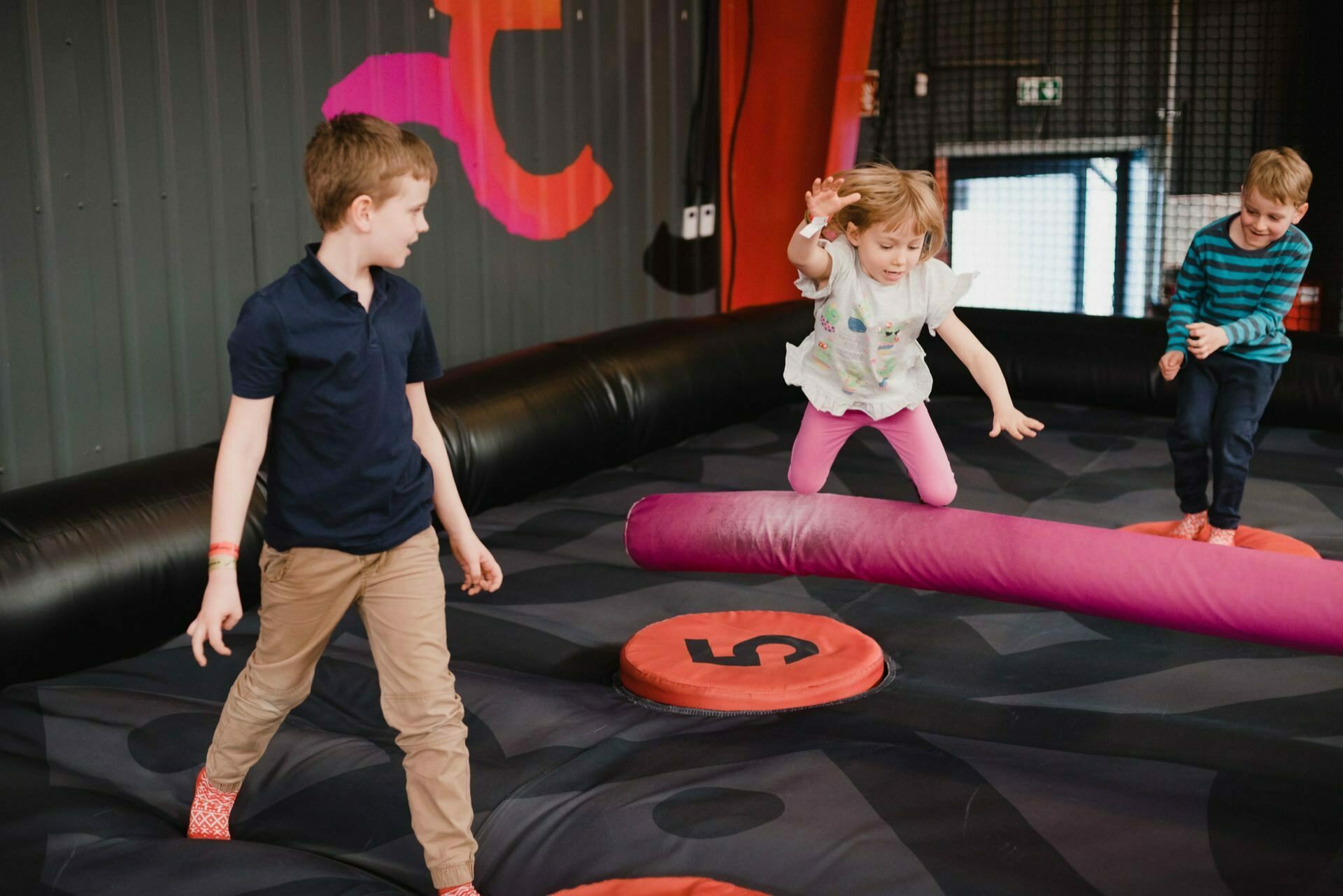 Three children are playing on an inflatable obstacle course indoors. One child in a white T-shirt jumps in the air over a pink cushioned beam, while two other children, one on the left and one on the right, watch and balance on the inflatable surface - perfect moments for event photography. 