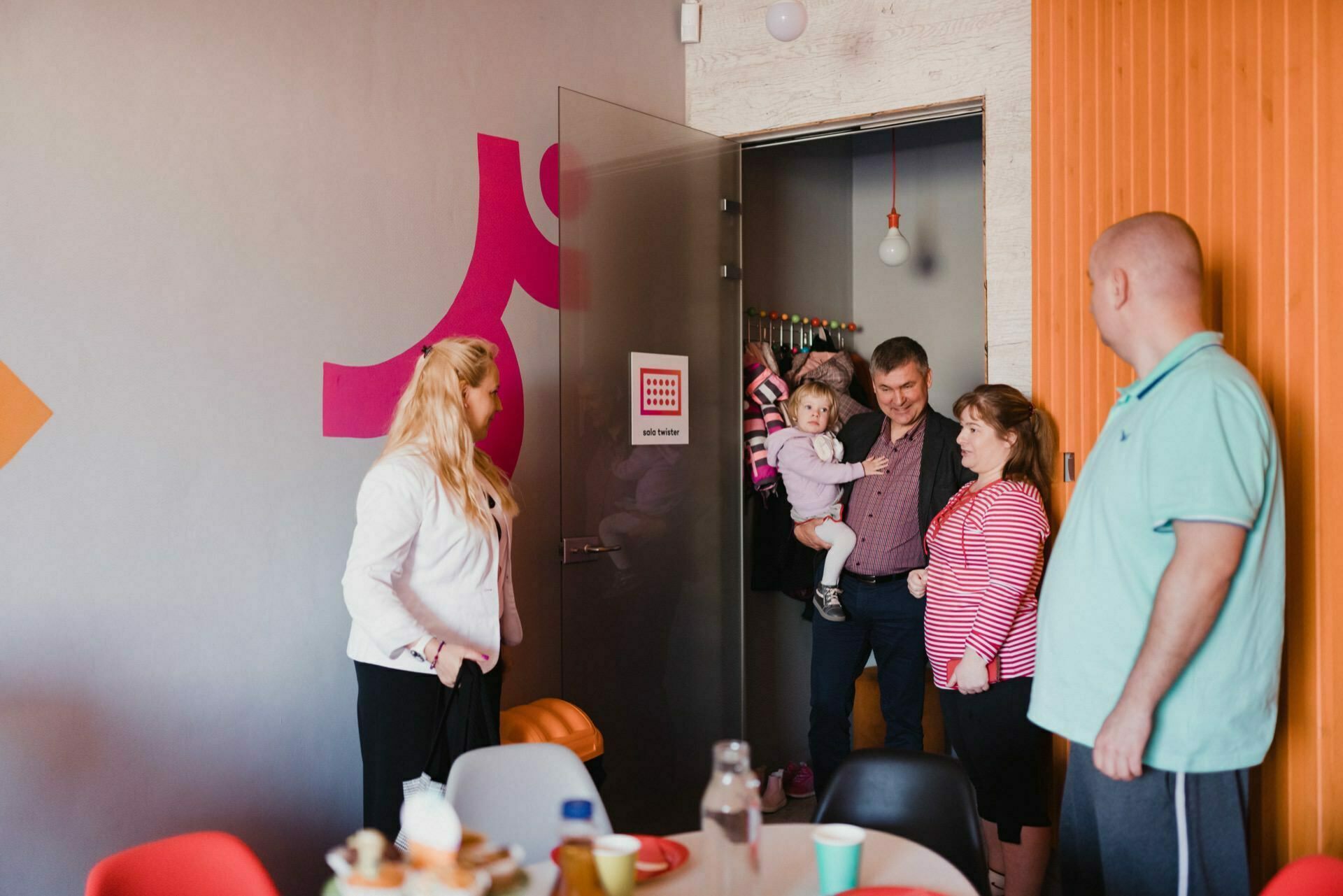 A man holding a small child enters a room with colorful decor. The three adults in the room, two women and a man, smile at them. The table is set for a meal with cups, a bottle and plates. In the background, bright pink and orange wall accents are visible - perfectly capturing this beautiful photo of the events.   