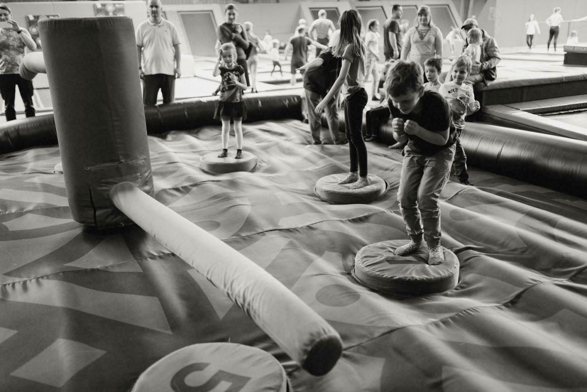 Children and adults play on an inflatable obstacle course. Some children balance on circular platforms while dodging a soft, rotating, padded beam. Spectators gather around the event, presumably captured in event photography. The mood is lively and fun.   