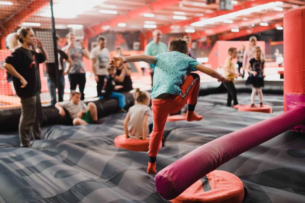 Dzieci i dorośli korzystają z krytego parku trampolin. Chłopiec w turkusowej koszuli i czerwonych spodniach przeskakuje nadmuchiwaną przeszkodę, podczas gdy inni obserwują i bawią się w tle. Przestrzeń jest wyposażona w miękkie, kolorowe konstrukcje do wspinaczki i skakania, idealne do żywej fotografii eventowa.