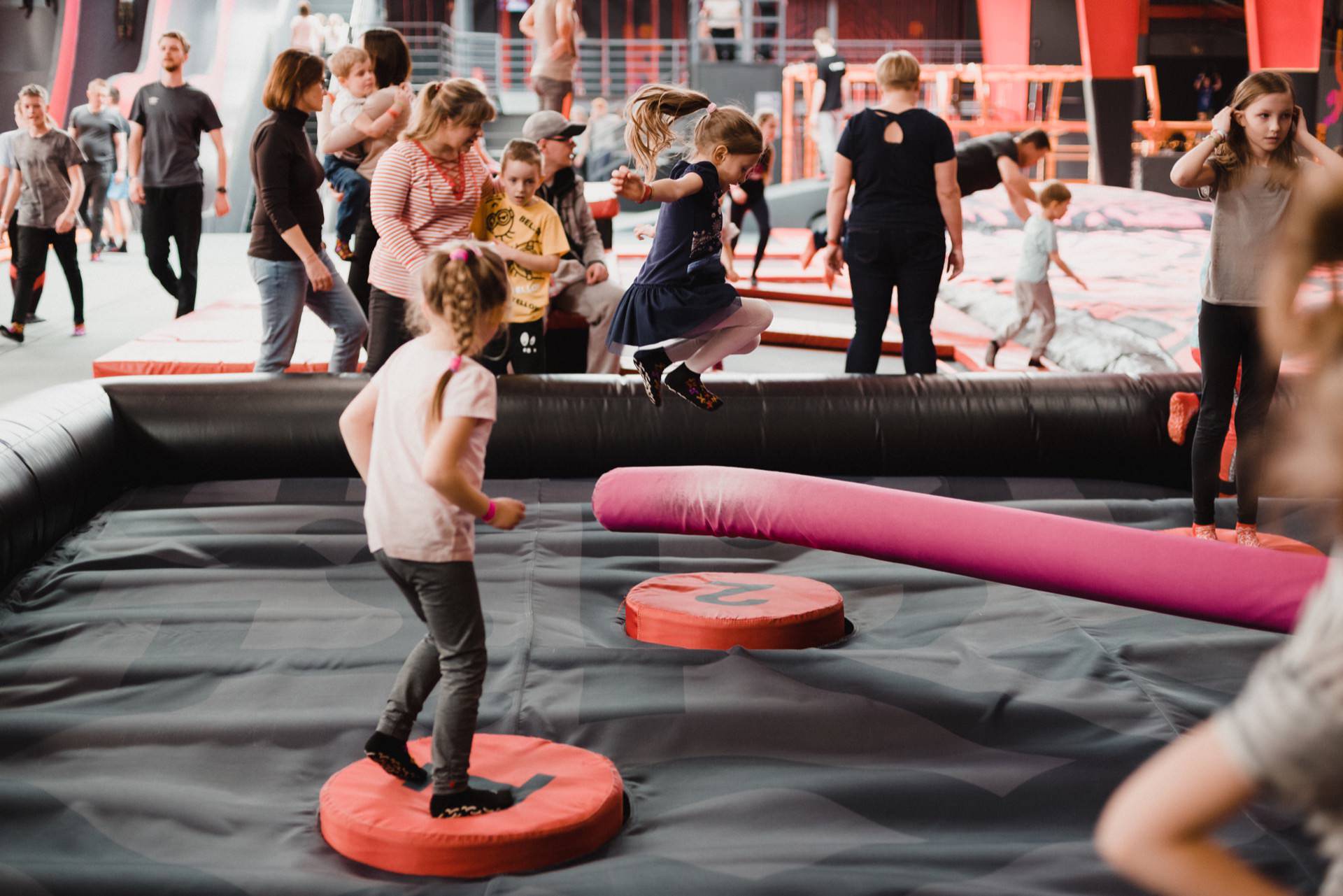 Children and adults can enjoy the trampoline park. Two girls stand on red round pads, while a third girl in a navy blue dress jumps high. Others interact and play in the background, and colorful equipment and padding can be seen throughout the space, creating an ideal location for event photography.  