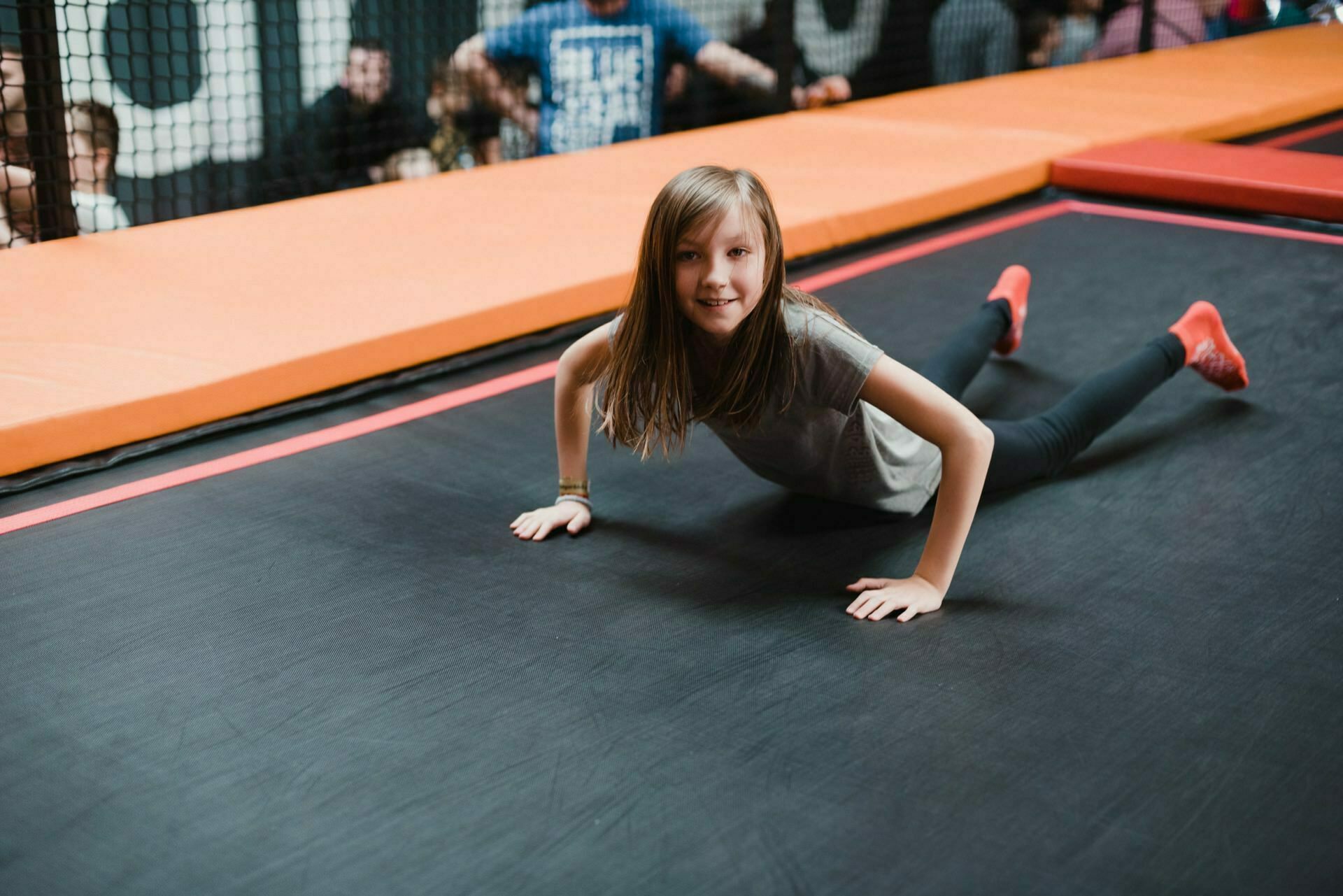 A girl with shoulder-length hair, wearing a gray t-shirt, black pants and red socks smiles while lying on her stomach on a trampoline. In the background of this event photo, a group of people can be seen behind a net. 