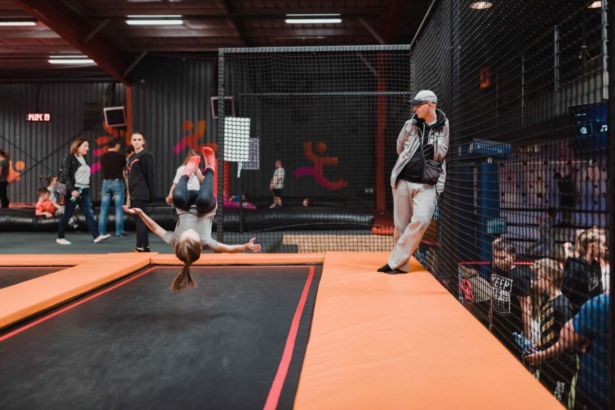 Młoda dziewczyna wykonuje przewroty na trampolinie w krytym parku trampolin, co doskonale uchwyciło to zdjęcie eventowe. Jest w powietrzu, z nogami nad głową, podczas gdy kobieta opiera się o płot z siatki i obserwuje. Inne dzieci i dorośli są obecni w tle, niektórzy patrzą, inni się bawią.