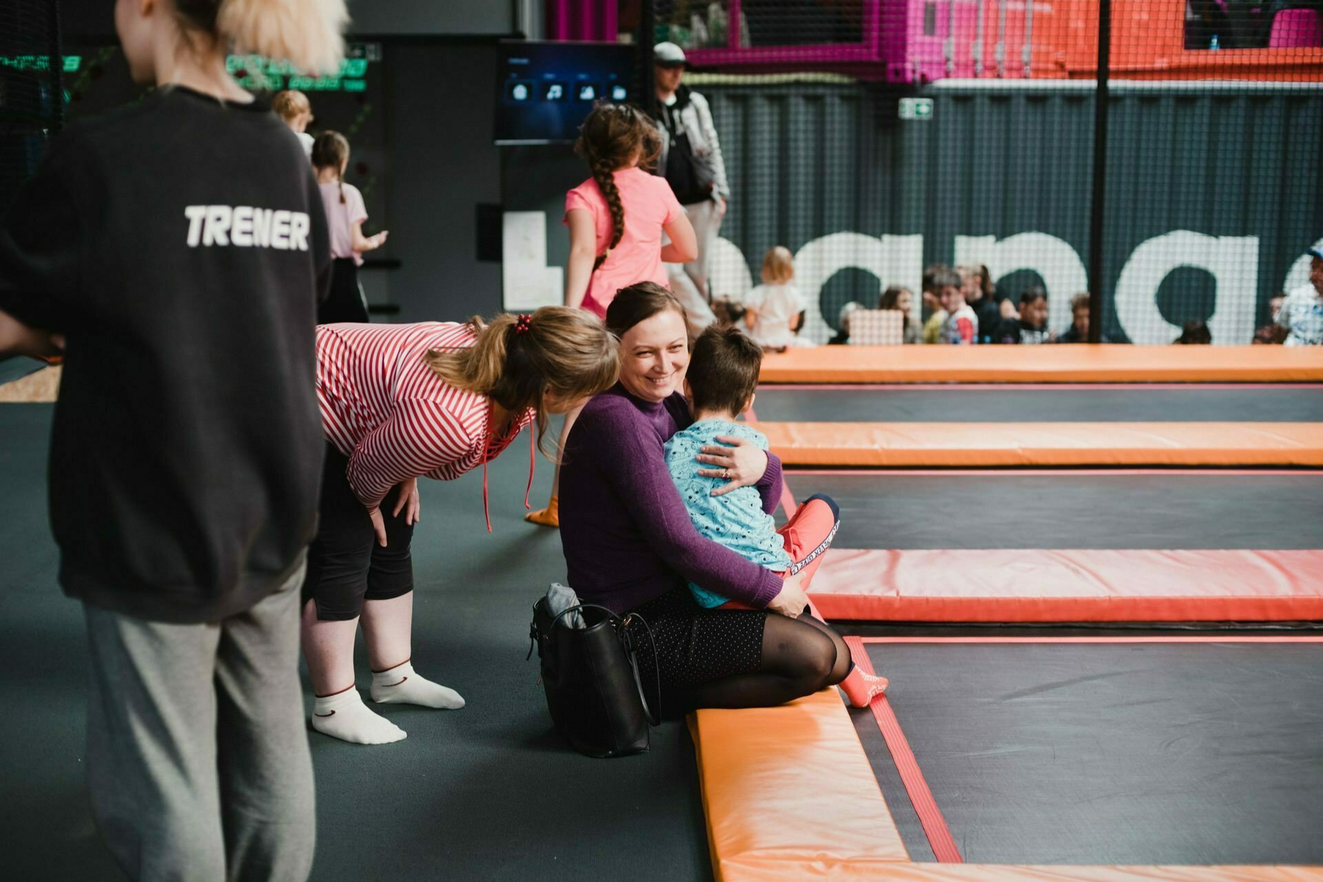 A woman in a purple sweater sits on the edge of a trampoline, holding a child on her lap. To talk to them, another woman in a red and white striped shirt and black shorts leans over. Several children are playing on nearby trampolines, all beautifully captured by event photography. A hangar sign can be seen in the background.   