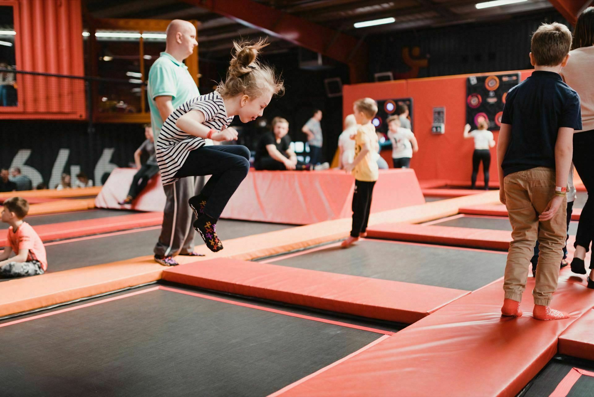 Children and adults are having fun at a trampoline park. A young girl in a striped shirt jumps in the air on a trampoline, while other children and adults are nearby on separate trampolines. The room is full of red and orange colors, perfectly captured in this event photo.  