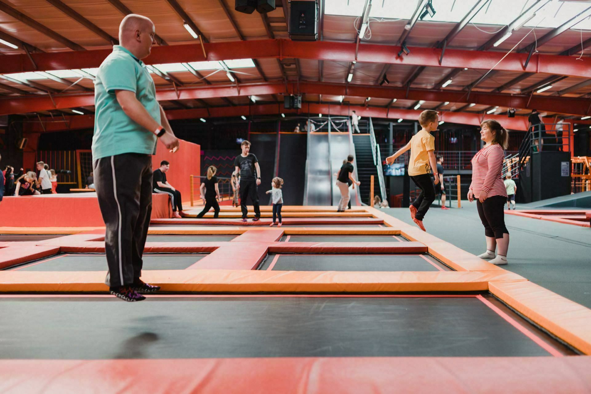 People are enjoying their time at an indoor trampoline park. A bald man in a green shirt jumps on a trampoline, while a boy and a girl jump in the air on neighboring trampolines. The other guests, including children and adults, are visible in the background, which was perfectly captured by the event photography.  