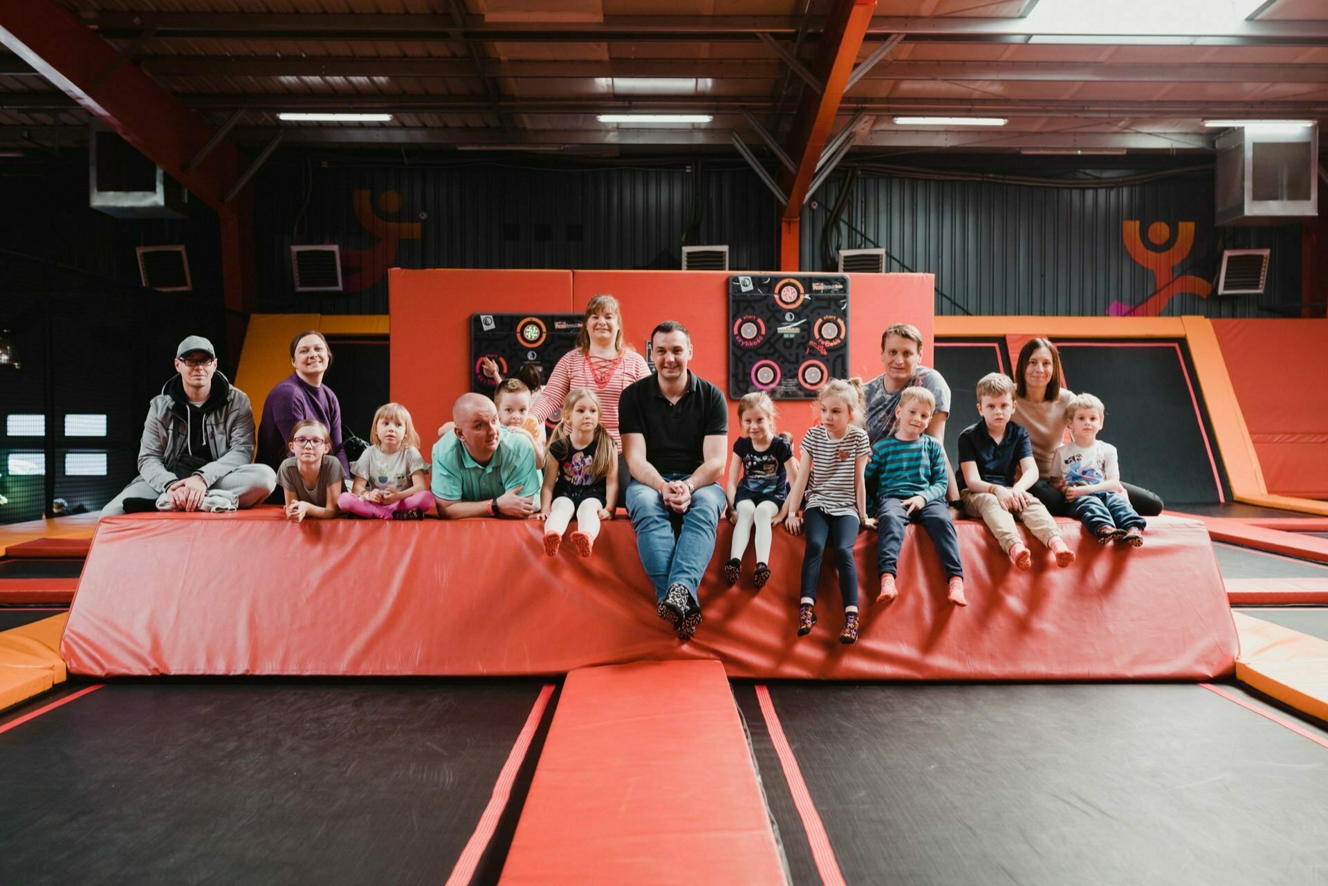 A group of adults and children sit together on the edge of a large trampoline in an indoor park. They smile and look into the camera, against a backdrop of colorful wall padding and play equipment. This vibrant photo report of the events perfectly captures the joyous moment.  