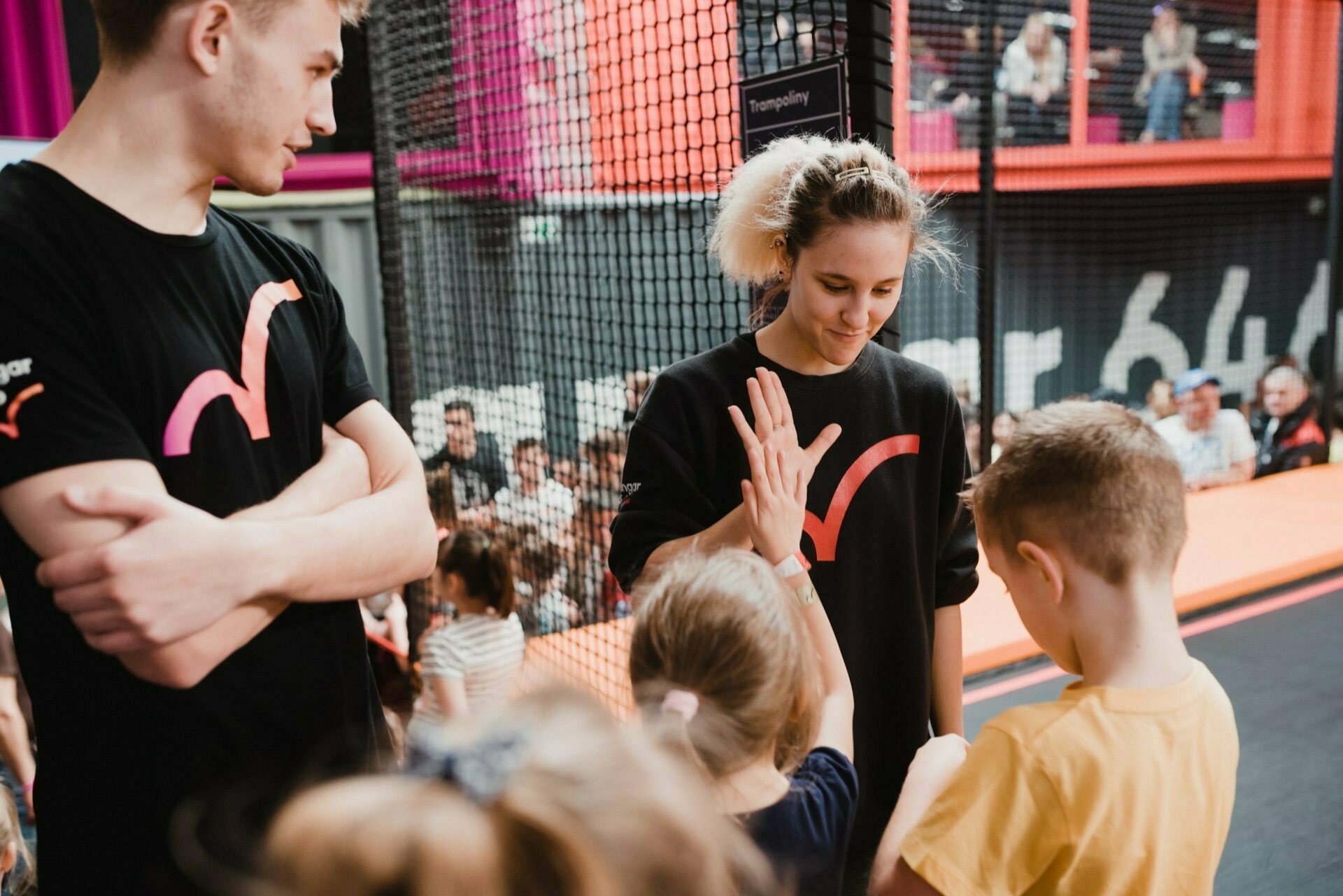 Two adults and two children interact at an indoor trampoline park in this delightful event photo essay. One adult is wearing a black shirt with an orange pattern, while the other high-fives a child in a yellow shirt. A trampoline net can be seen in the background with spectators enjoying the fun.  