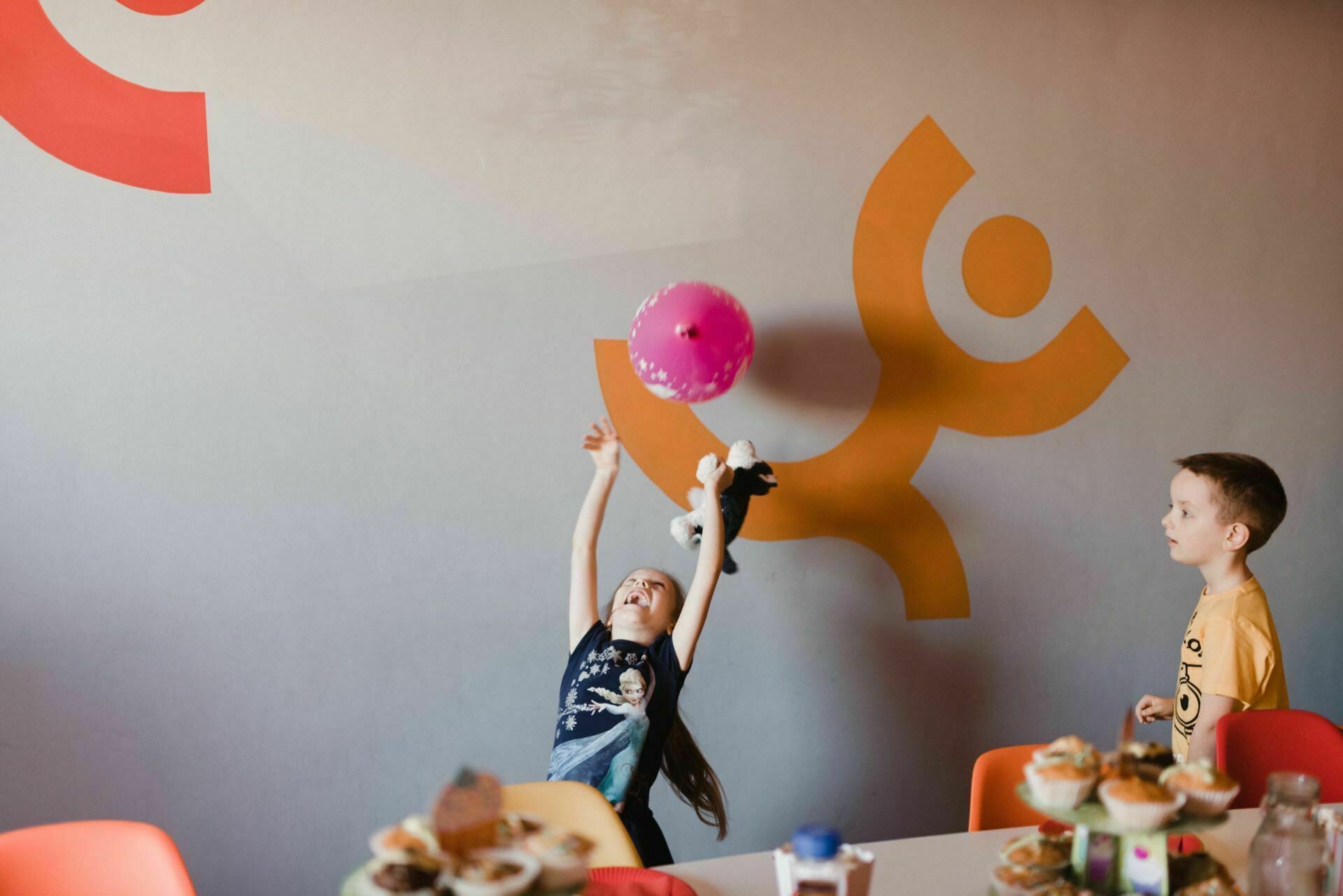 A young girl reaches out to grab a pink balloon, while a boy stands nearby and watches. The room captured in this event photo has colorful wall decorations, including yellow and orange human figures. Tables with food and drinks are surrounded by orange chairs, as beautifully documented in the event photo.  