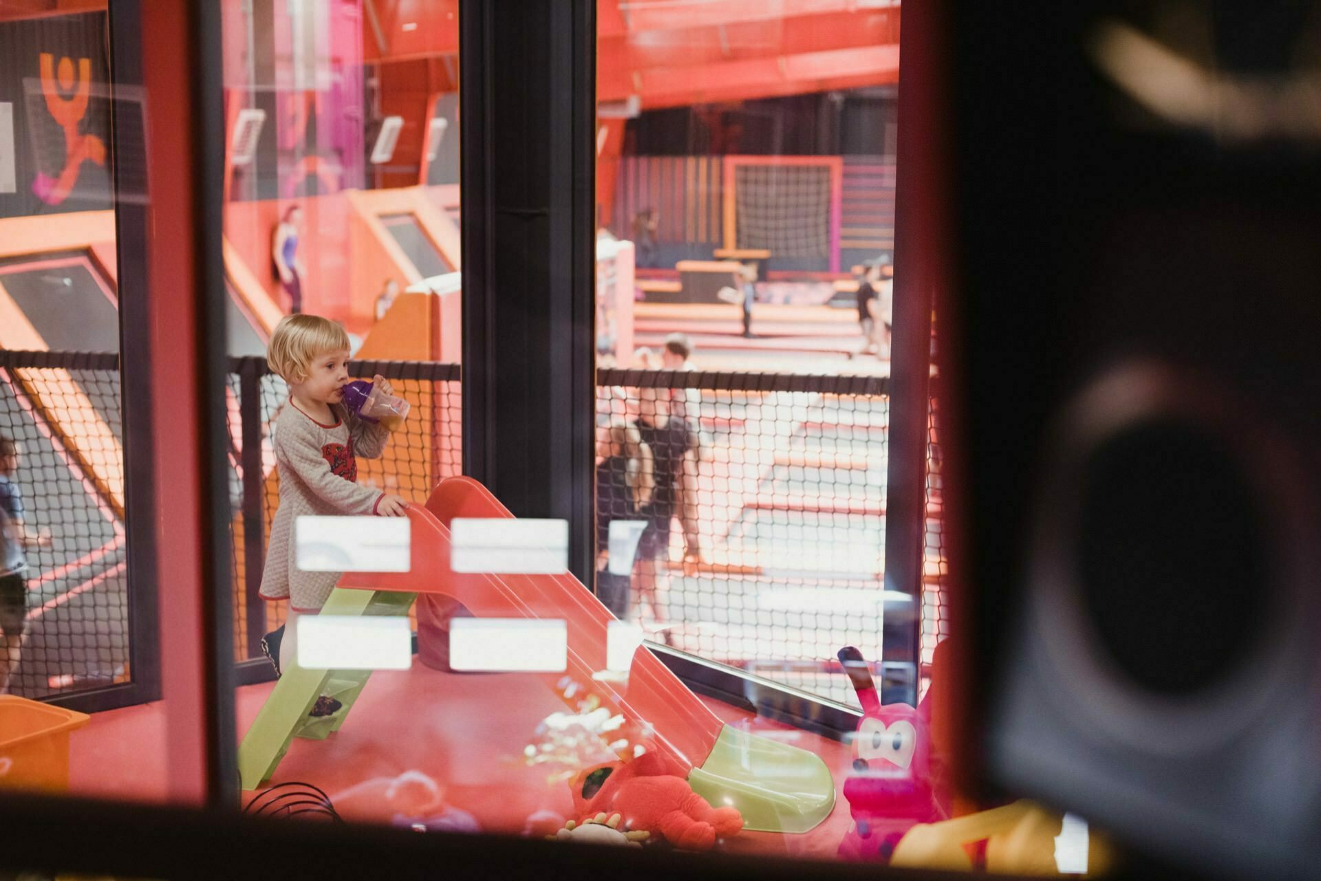 A young child stands at the top of a small slide in an indoor playground holding a drink. In the background you can see a trampoline park with people jumping on trampolines. Bright and colorful toys can be seen near the slide, capturing the essence of event photography and event photojournalism.  