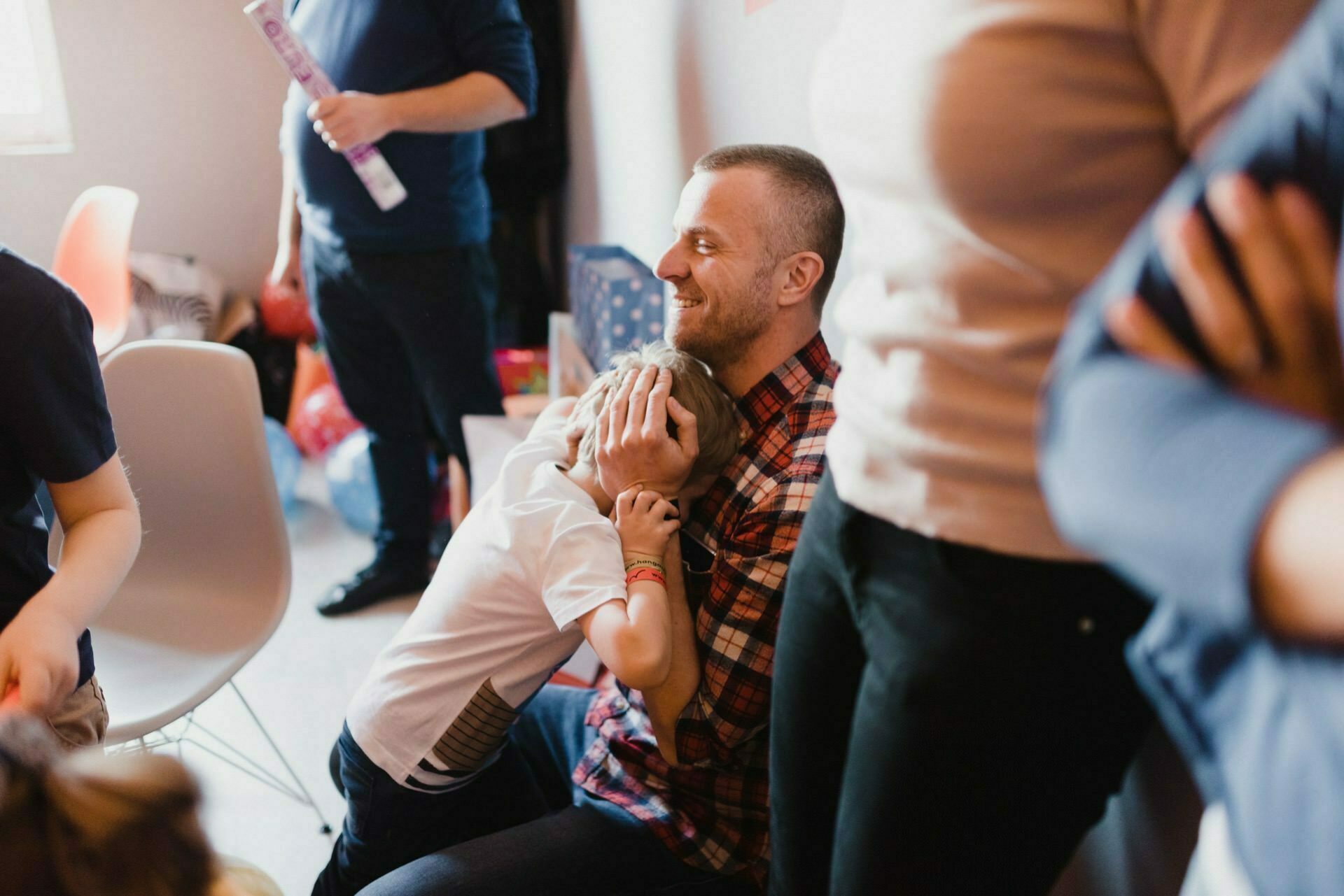 A man in a checked shirt kneels on the floor, smiling and hugging a small child who is burying his face in his chest. People stand or sit around them in the brightly lit room, beautifully captured as part of the photo coverage of the events. 