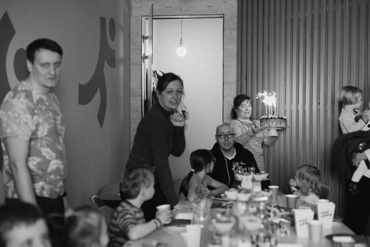 Grupa ludzi, w tym dorośli i dzieci, gromadzi się wokół stołu, aby świętować. Stół zastawiony jest filiżankami i przekąskami. W tle przykuwa uwagę kobieta niosąca tort z zapalonymi świeczkami. Scena wydaje się radosna i odświętna – idealna na fotografię eventową w Warszawie.