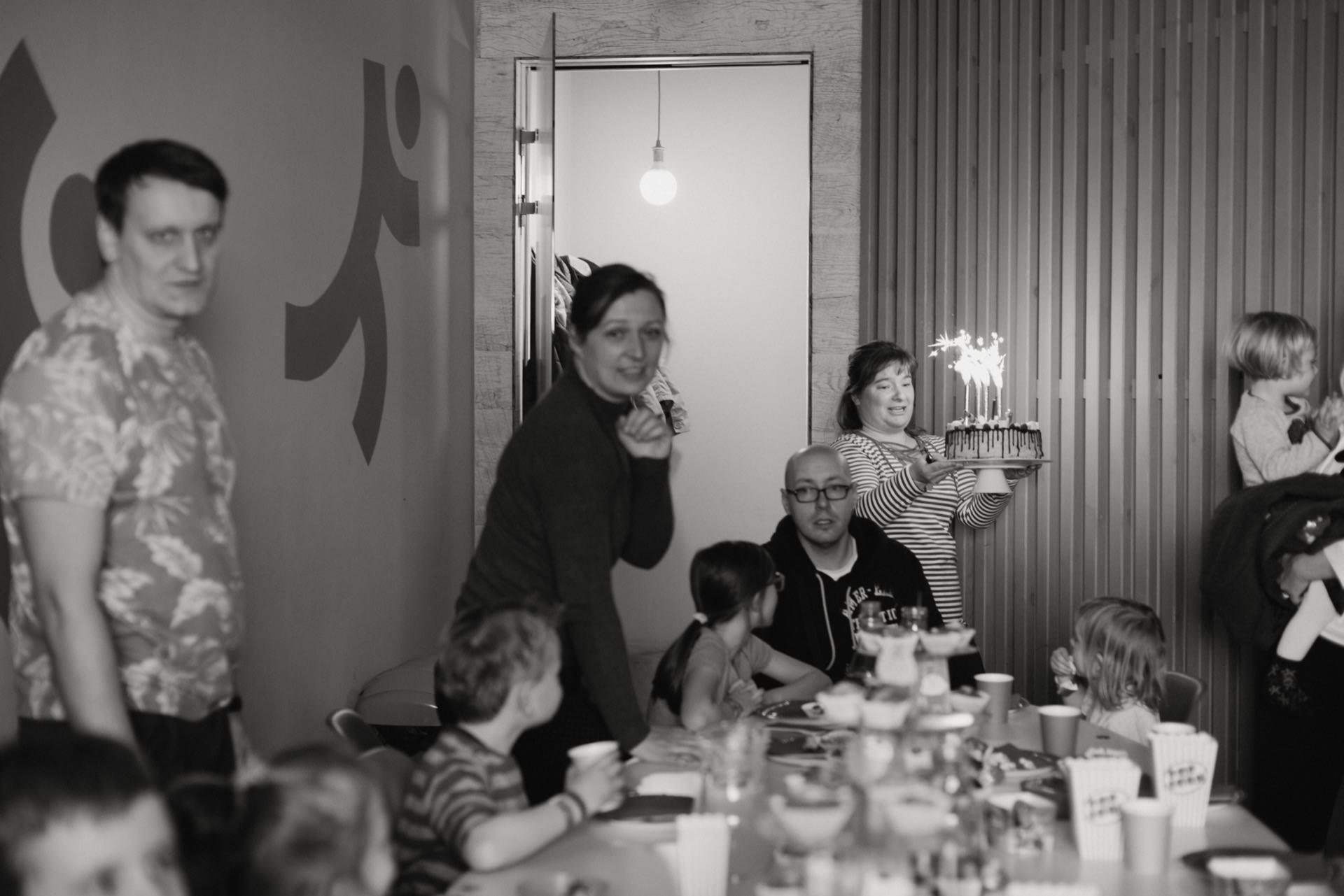 A group of people, including adults and children, gather around a table to celebrate. The table is set with cups and snacks. A woman carrying a cake with lit candles catches the eye in the background. The scene seems joyful and festive - perfect for event photography in Warsaw.   