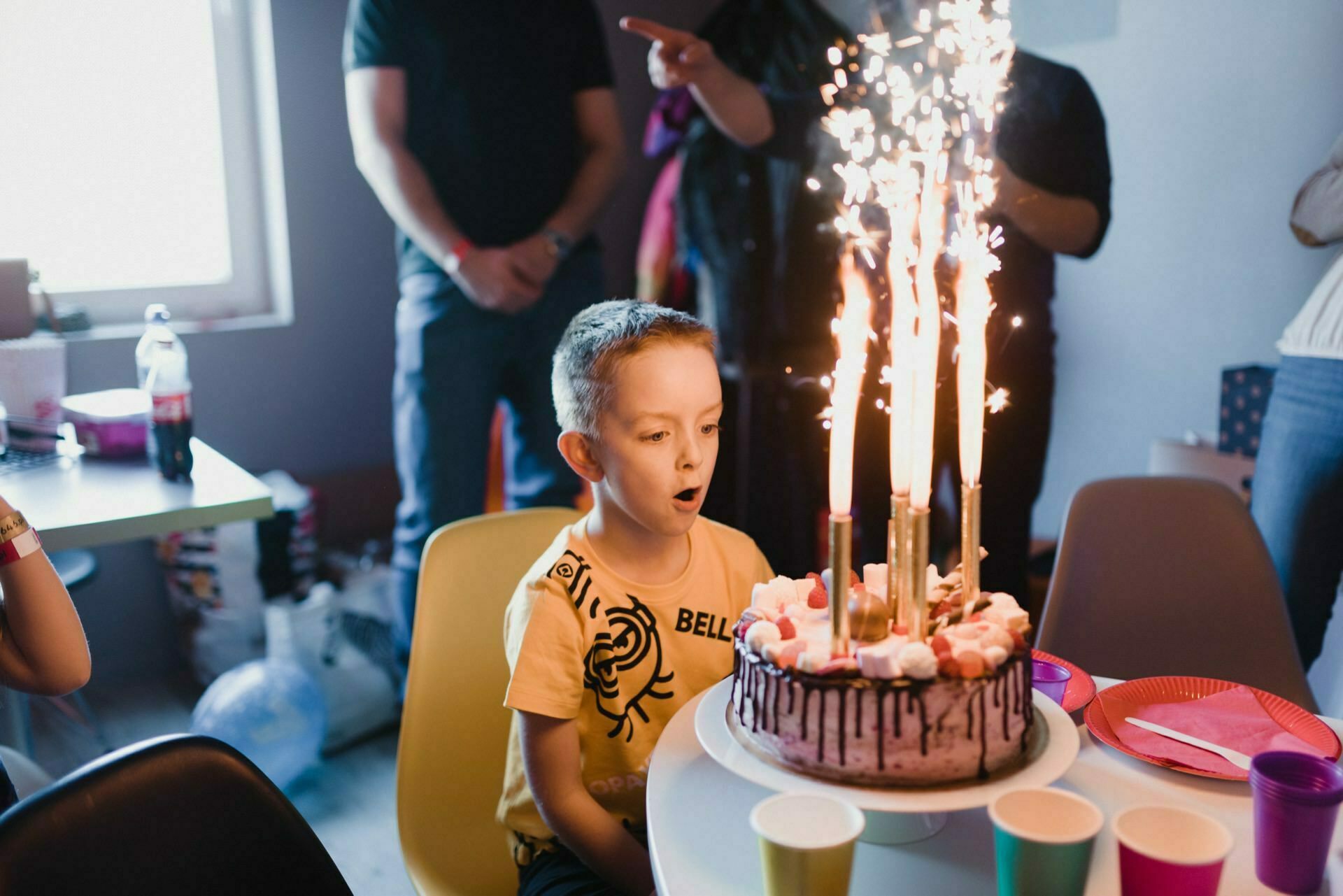 A young boy looks excitedly at a birthday cake decorated with large sparkling candles. Other people are standing around him, and the room is decorated for the celebration. Bright teacups are arranged on the table, perfectly capturing the essence of this joyful moment in this event photography.  