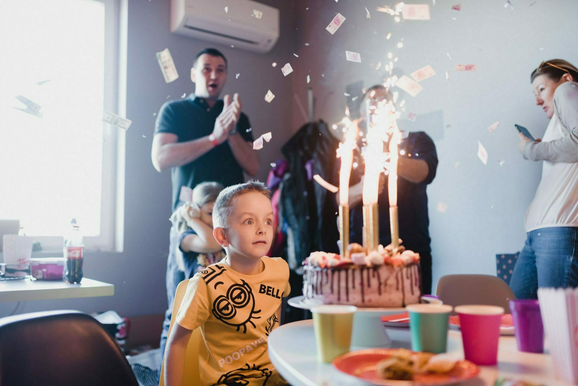 A young boy sits at a table with his eyes wide open and stares at a birthday cake decorated with tall sparklers. Behind him, an adult claps his hands, while another captures a photo moment of the events. Confetti floats in the air, colorful cups and plates stand on the table, and an air conditioner is mounted on the wall.  